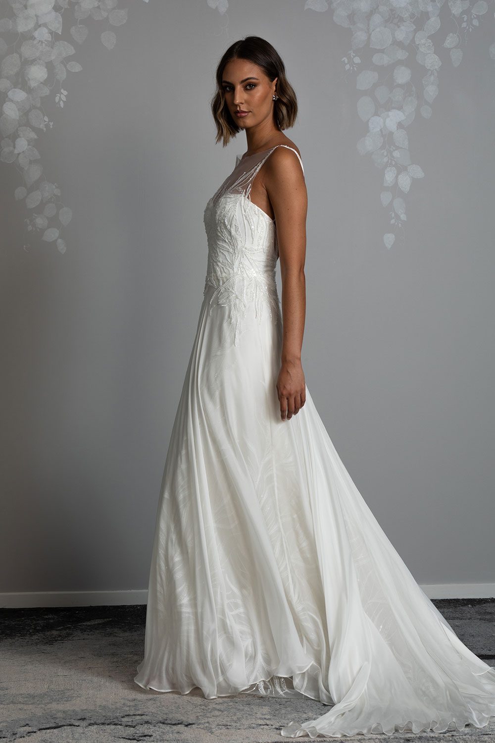 Gio Wedding Dress from Vinka design. Intricate gown with high sheer neckline and a fitted, structured bodice, as well as a double-strapped low back. The lace is hand-appliqued throughout the bodice and flatters the smallness of the waist, providing a stunning silhouette. Model in profile view showing long silk chiffon train that moves beautifully