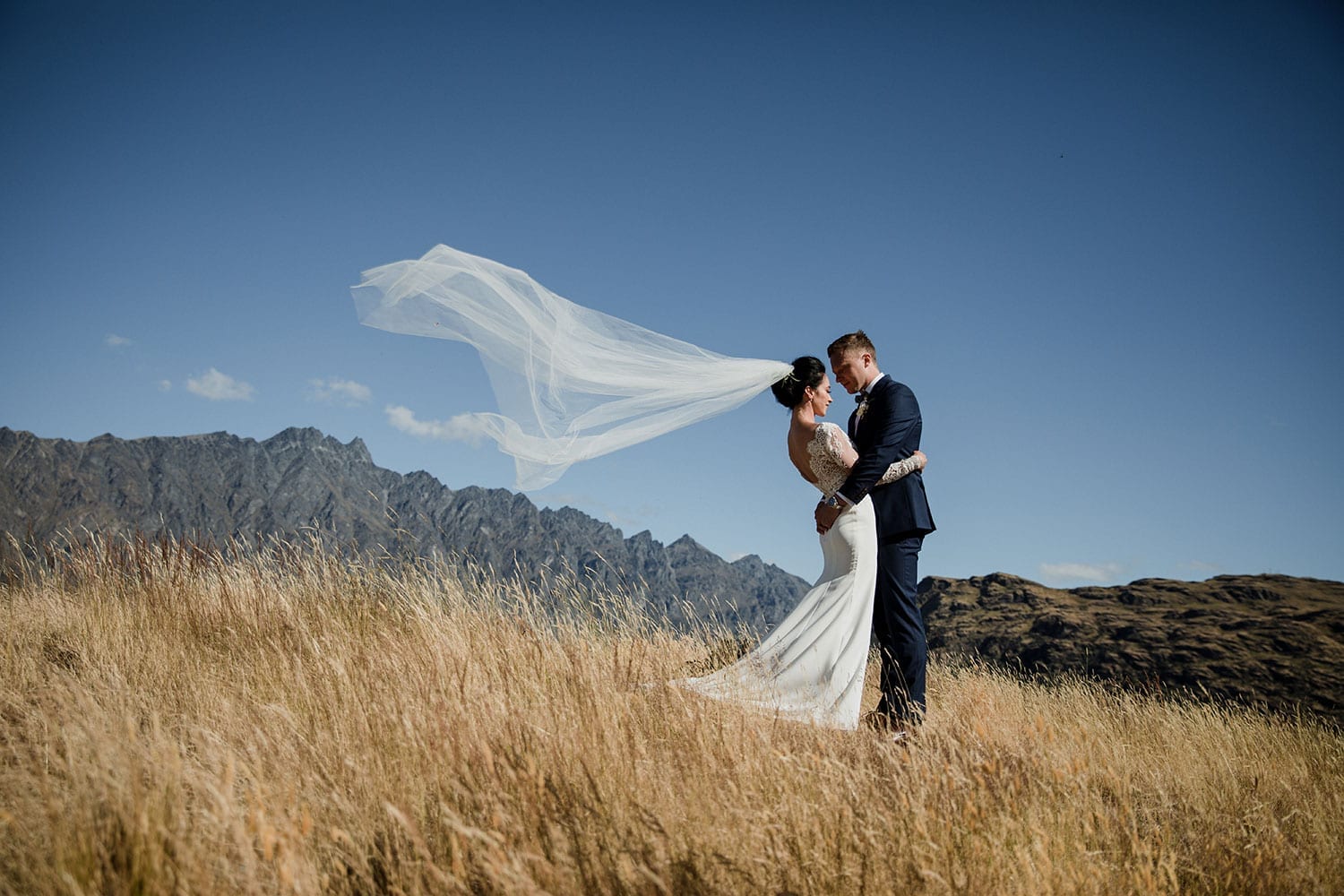 Vinka Design Features Real Weddings - bride in bespoke custom made gown with groom under veil in breeze with Queenstown with mountains in background
