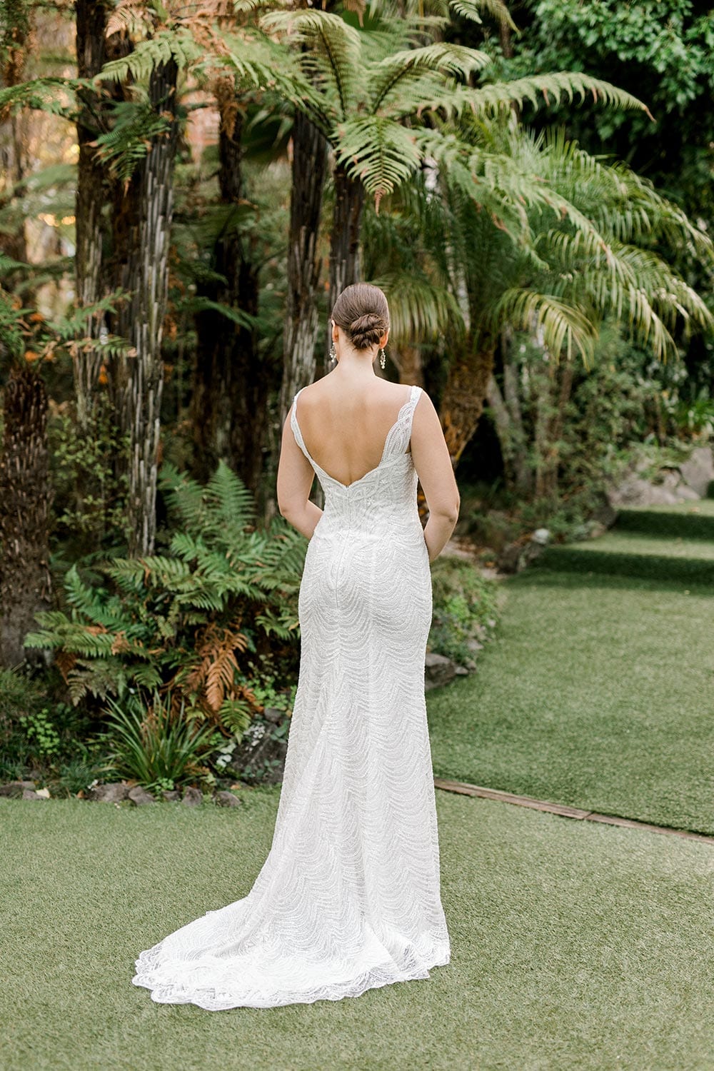 Juliette Wedding Dress from Vinka Design. Flattering stretch fitted lace wedding dress with beautiful ivory rich beading. Structured bodice provides support while remaining effortless to wear. Full length of dress from behind with train to the side. Photographed at Tui Hills.