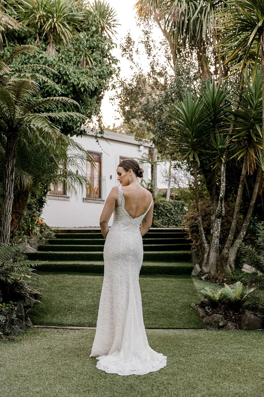 Juliette Wedding Dress from Vinka Design. Flattering stretch fitted lace wedding dress with beautiful ivory rich beading. Structured bodice provides support while remaining effortless to wear. Full length of dress from behind. Photographed at Tui Hills.
