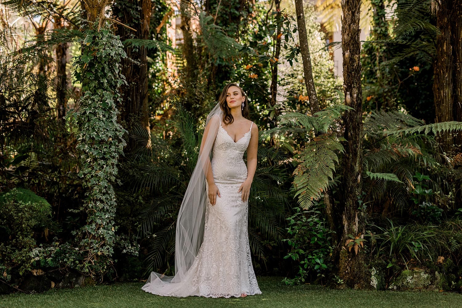 Sahara Wedding Dress from Vinka Design. Stunning fitted lace wedding gown. Lightly-beaded floral lace highlights the ivory stretch satin base. Sweetheart neckline & low back with beautiful lace detail. Full length of dress. Photographed in landscaped gardens.