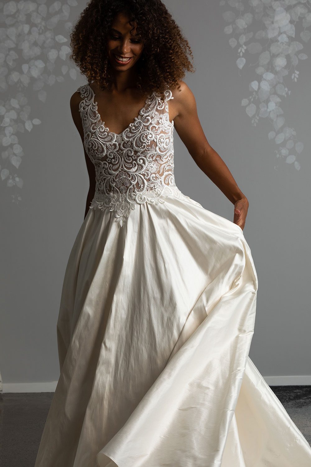 Brooke Wedding Dress from Vinka Design. This stunning wedding dress has a structured bodice intricately layered to emphasize natural curves. Sweetheart neckline, delicate lace-embellished straps, low back & silk chiffon train. Front view of model holding dupion silk skirt