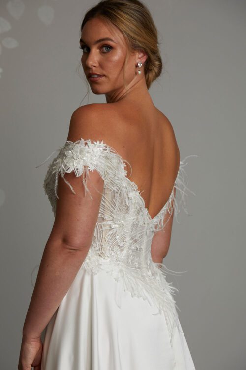 Genevive Wedding Dress from Vinka Design. The soft off-the-shoulder sleeves & semi-sheer structured bodice with hand-appliqued lace and a soft satin skirt make this a romantic & dreamy wedding dress. Close up of back of dress with deep V and off shoulder lace straps with feather details