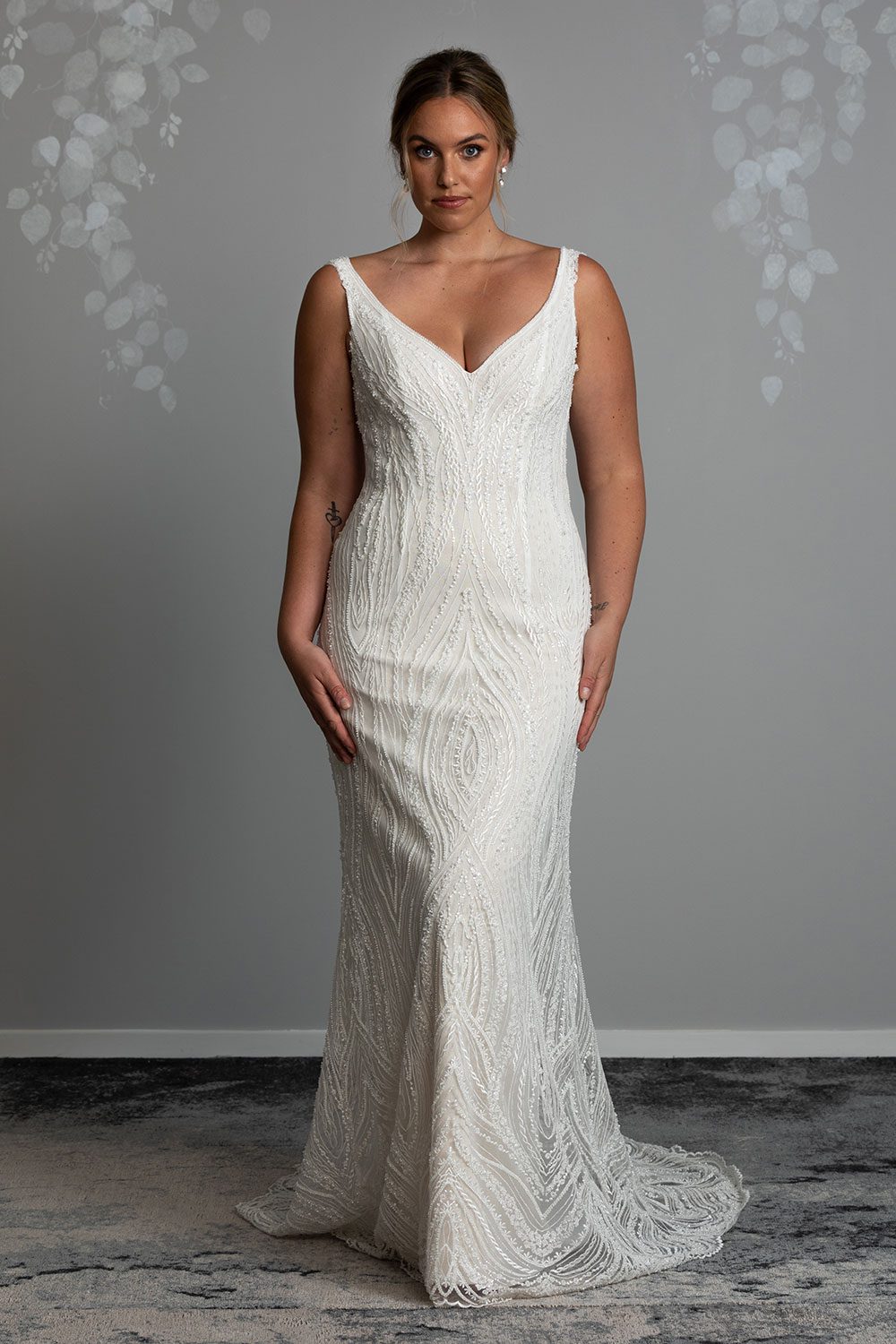 Kiera Wedding Dress from Vinka Design. Beautifully beaded lace wedding dress. Deep V-neckline both front and back is complemented with delicate sheer lace shoulder detail & structured bodice. Model with hands to hips wearing deep V-neckline dress with beaded lace