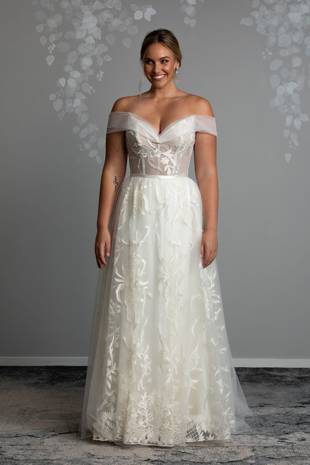 Zoe wedding dress by Vinka Design - a romantic & dreamy off-shoulder gown with a structured, boned bodice, a full lace & tulle skirt. Semi-sheer bodice with tulle off-shoulder detailing and satin belt. Model wearing off-shoulder gown with boned bodice and full lace and tulle skirt