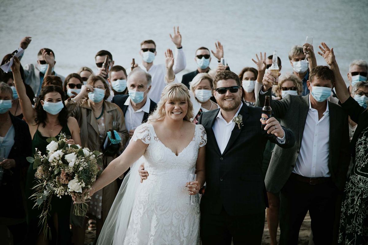 Vinka Design Features Real Weddings - bride in bespoke wedding gown posing with groom and wedding guests at the beach.