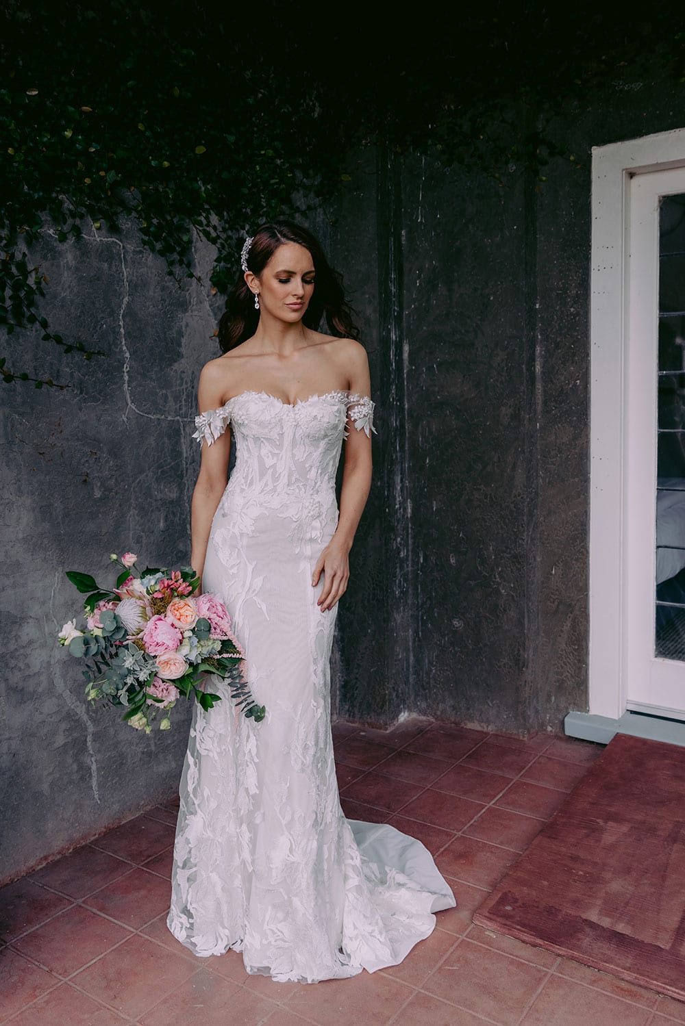 Sachi Wedding gown from Vinka Design - This spectacular wedding dress is off-shoulder and form-fitting to show off your figure to perfection. Richly embroidered lace is highlighted with small three-dimensional leaves and flowers. Model wearing gown showing full length of dress.