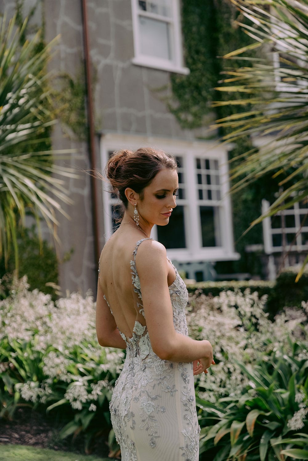 Kazumi Wedding gown from Vinka Design - Flattering and form fitting wedding dress in soft grey shades on a blush base. Low sheer back is adorned with hand-appliqued lace, bodice has a deep V-neckline, & thin straps that compliment the shoulders. Model wearing gown walking through Clevedon gardens showing detail of low back of dress.