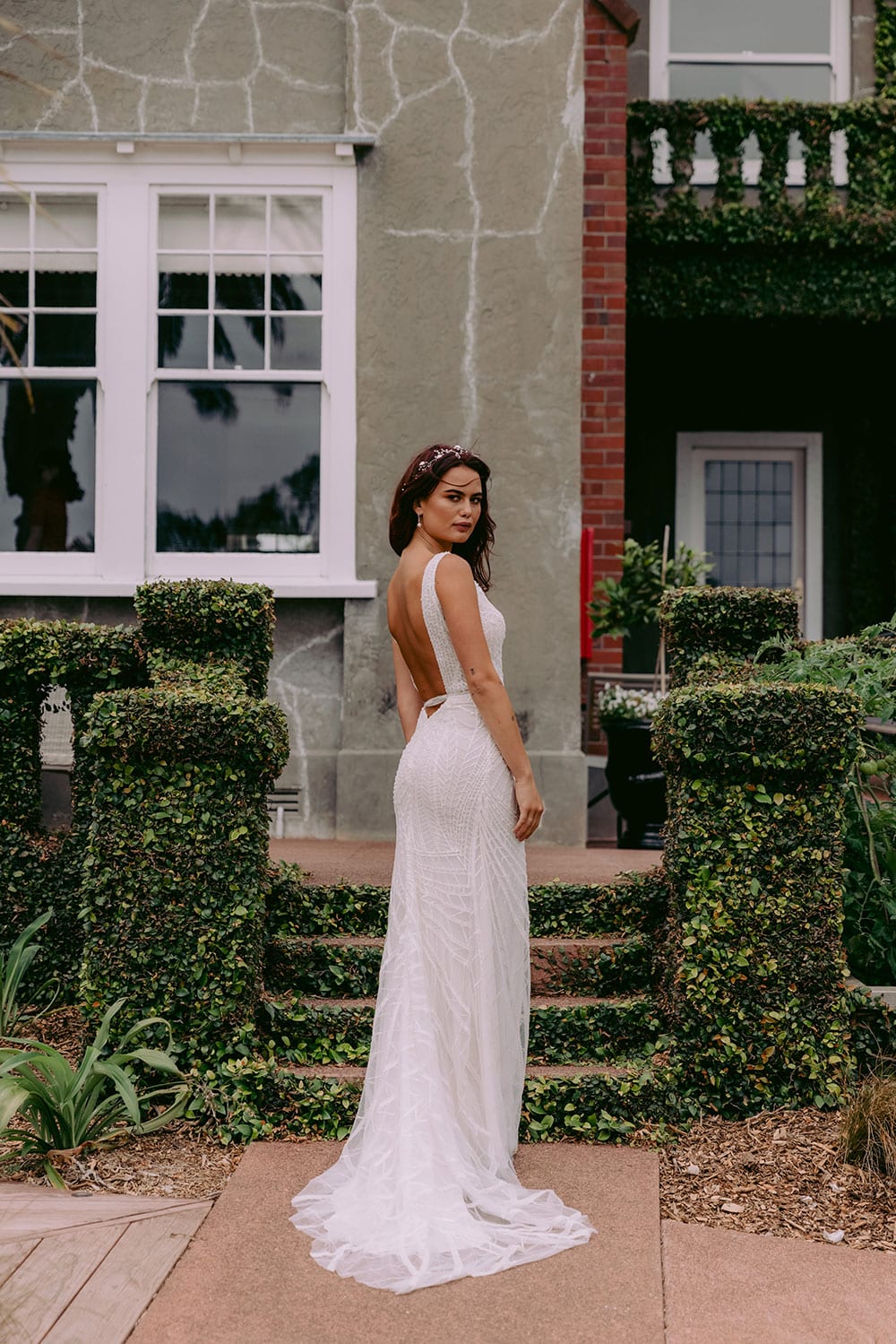 Elle Wedding gown from Vinka Design - Wedding dress with illusion neckline and deep plunge in the front with low back. Stunning beaded embroidery over a stretch base that sculpts and flatters the figure and falls into a beautiful flared train. Model wearing gown in beautiful gardens showing dress low back.
