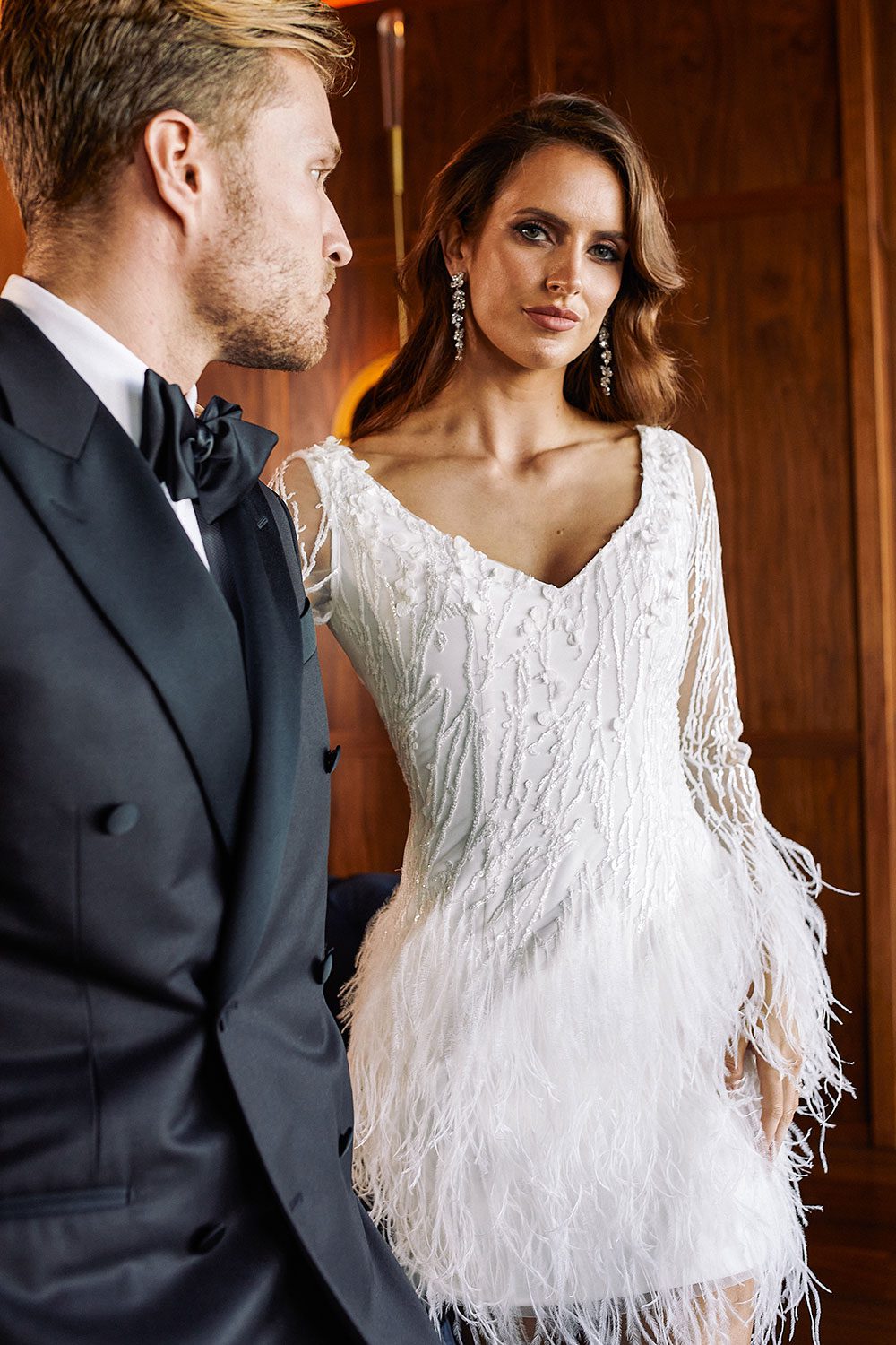 Nikora Wedding gown from Vinka Design - This sexy gown is sure to turn heads! Adorned with feathers that accentuate movement and bell sleeves that add flare, a high neckline, and low back. Close up of bride standing next to groom in a hotel