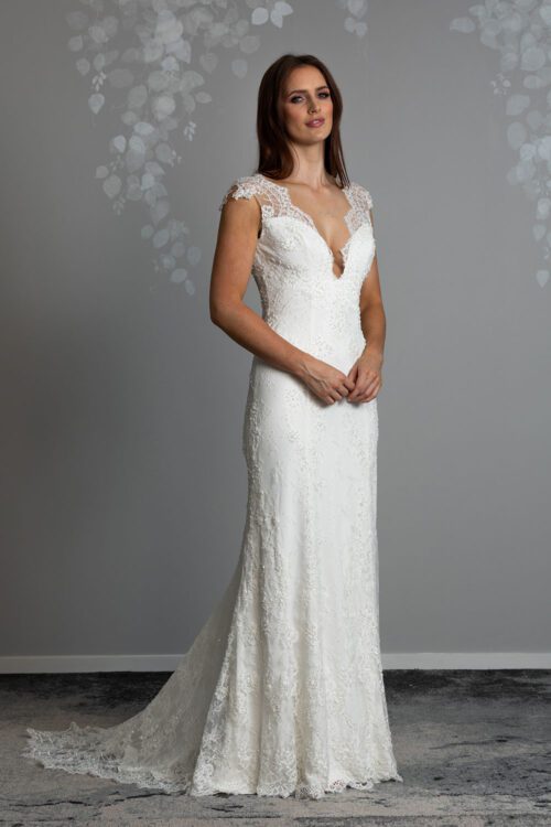 Fallyn Wedding gown from Vinka Design - This wedding dress is a timeless classic. Dramatic scallop lace and deep V-shaped neckline with beautiful low back and mini cap sleeves. This gown is cut in a fit-and-flare design with a side split. Model with hands clasped at front showing the deep v neckline with scalloped lace and long fit and flare skirt