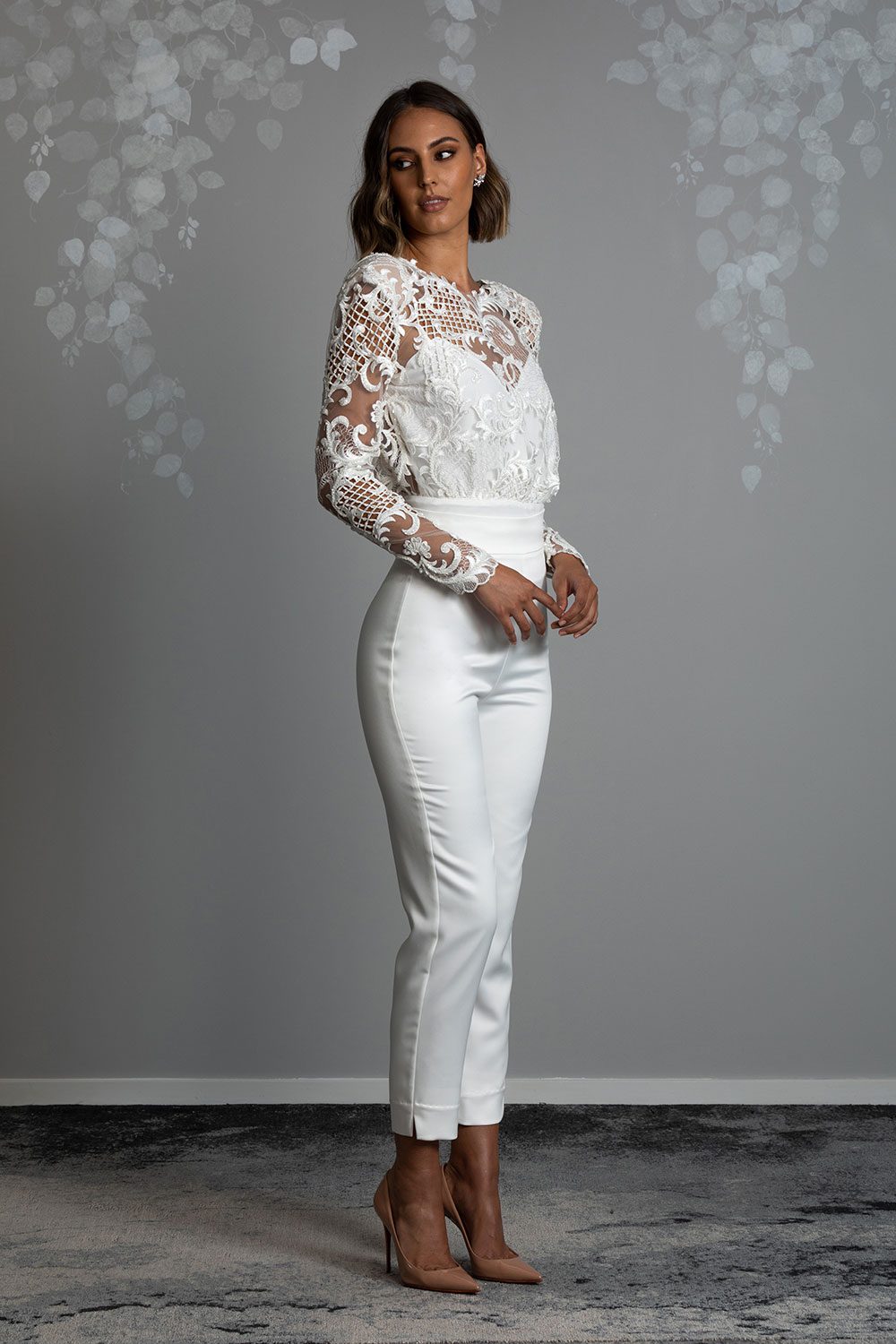 Ava Wedding Trousers and Blouse from Vinka Design - Elegantly tailored trouser combined with a beautiful lace blouse for the Modern Bride. The blouse features a high neck and low back, with in-built camisole, fitted sleeves, & pearl buttons. Model showing of semi profile view of form fitting pants with sheer embroidered lace blouse with long sleeves