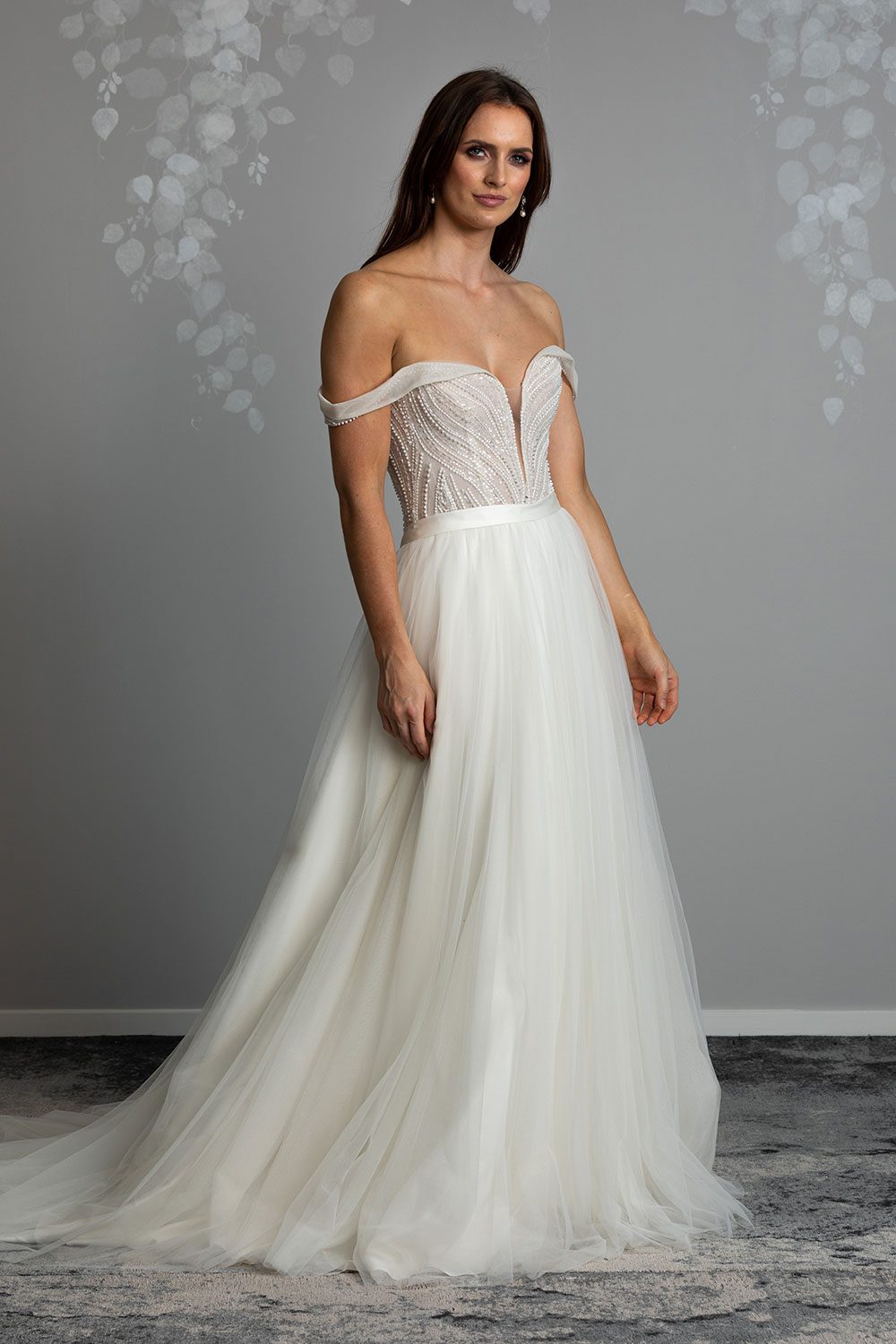 Aroha Wedding gown from Vinka Design - Beautiful & intricate pearl beading is hand-appliqued along this wedding gown’s structured, boned bodice. Delicate off-shoulder straps are complemented by the sweetheart neckline. - Model showing semi profile of sinched waist with satin belt and soft tulle skirt