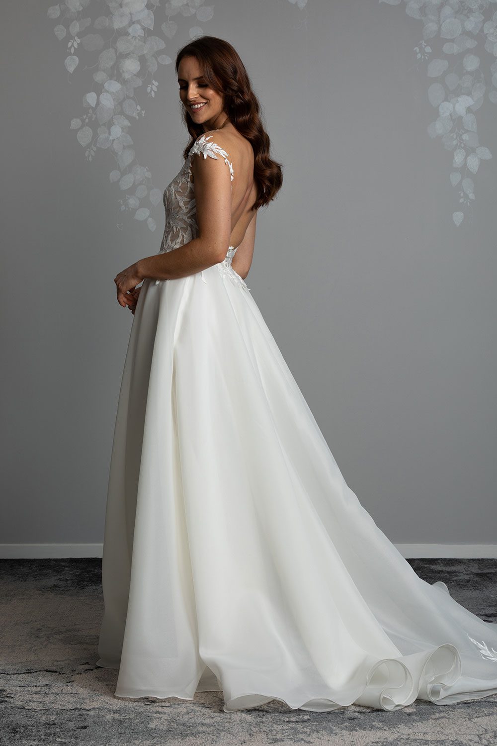 Ariana Wedding gown from Vinka Design - This gorgeous gown features delicate leaf lace hand-appliqued throughout the semi-sheer, structured bodice and up over the shoulder and skirt made of dreamy satin organza layers. Profile view of model showing off the long flowing train and low back with semi sheer lace detailing