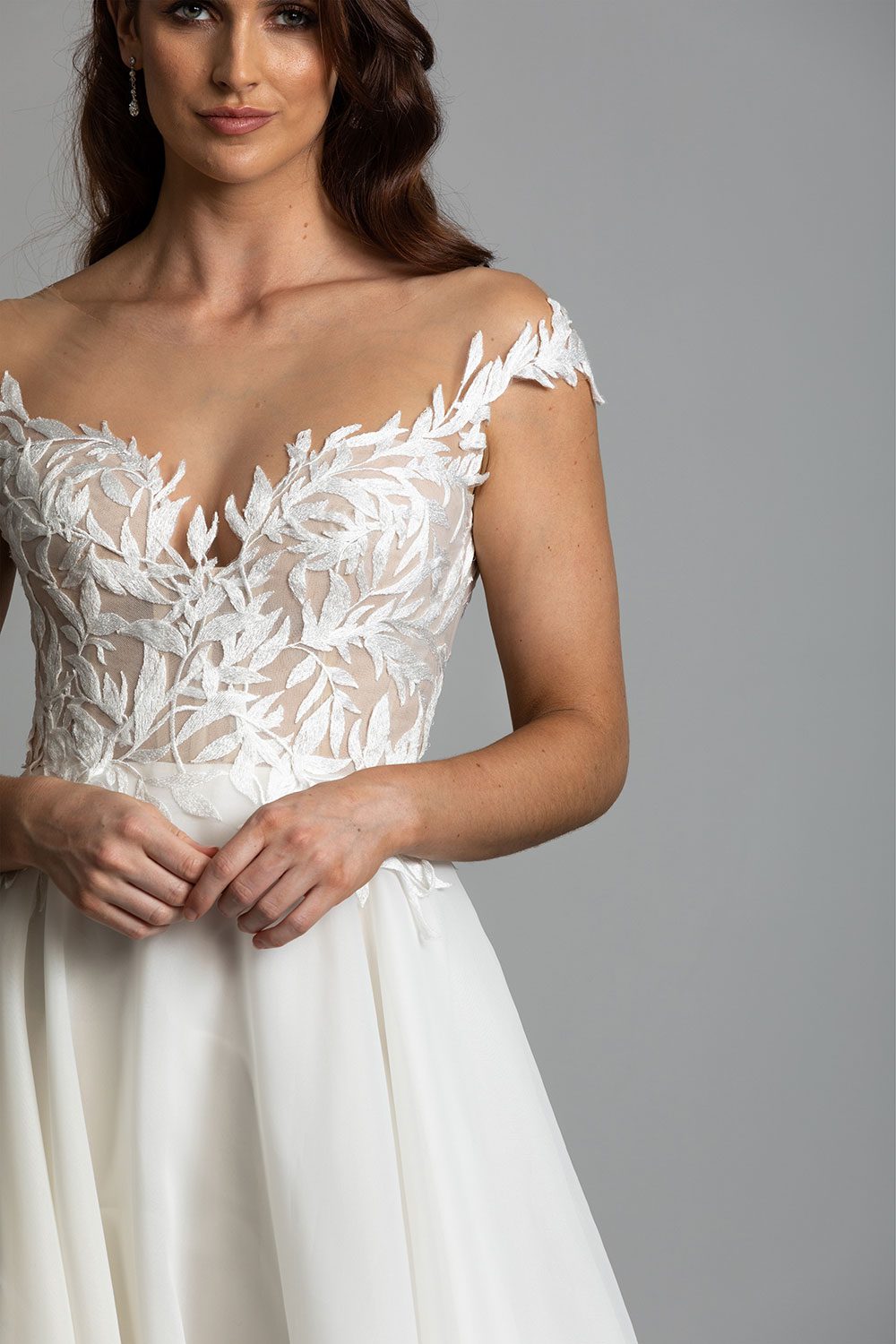 Ariana Wedding gown from Vinka Design - This gorgeous gown features delicate leaf lace hand-appliqued throughout the semi-sheer, structured bodice and up over the shoulder and skirt made of dreamy satin organza layers. Close up of model displaying semi-sheer bodice with off shoulder ivy lace details