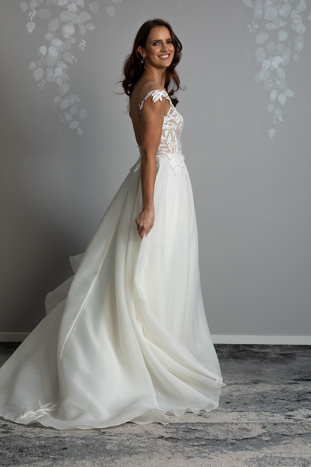 Ariana Wedding gown from Vinka Design - This gorgeous gown features delicate leaf lace hand-appliqued throughout the semi-sheer, structured bodice and up over the shoulder and skirt made of dreamy satin organza layers. Profile view showing translucent flowing organza skirt against low back and off shoulder ivy lace