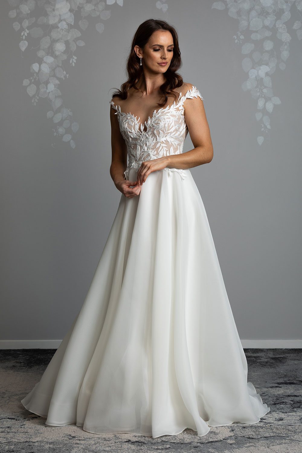 Ariana Wedding gown from Vinka Design - This gorgeous gown features delicate leaf lace hand-appliqued throughout the semi-sheer, structured bodice and up over the shoulder and skirt made of dreamy satin organza layers. Front view of model with hands clasped across waist where the bodice and skirt meet