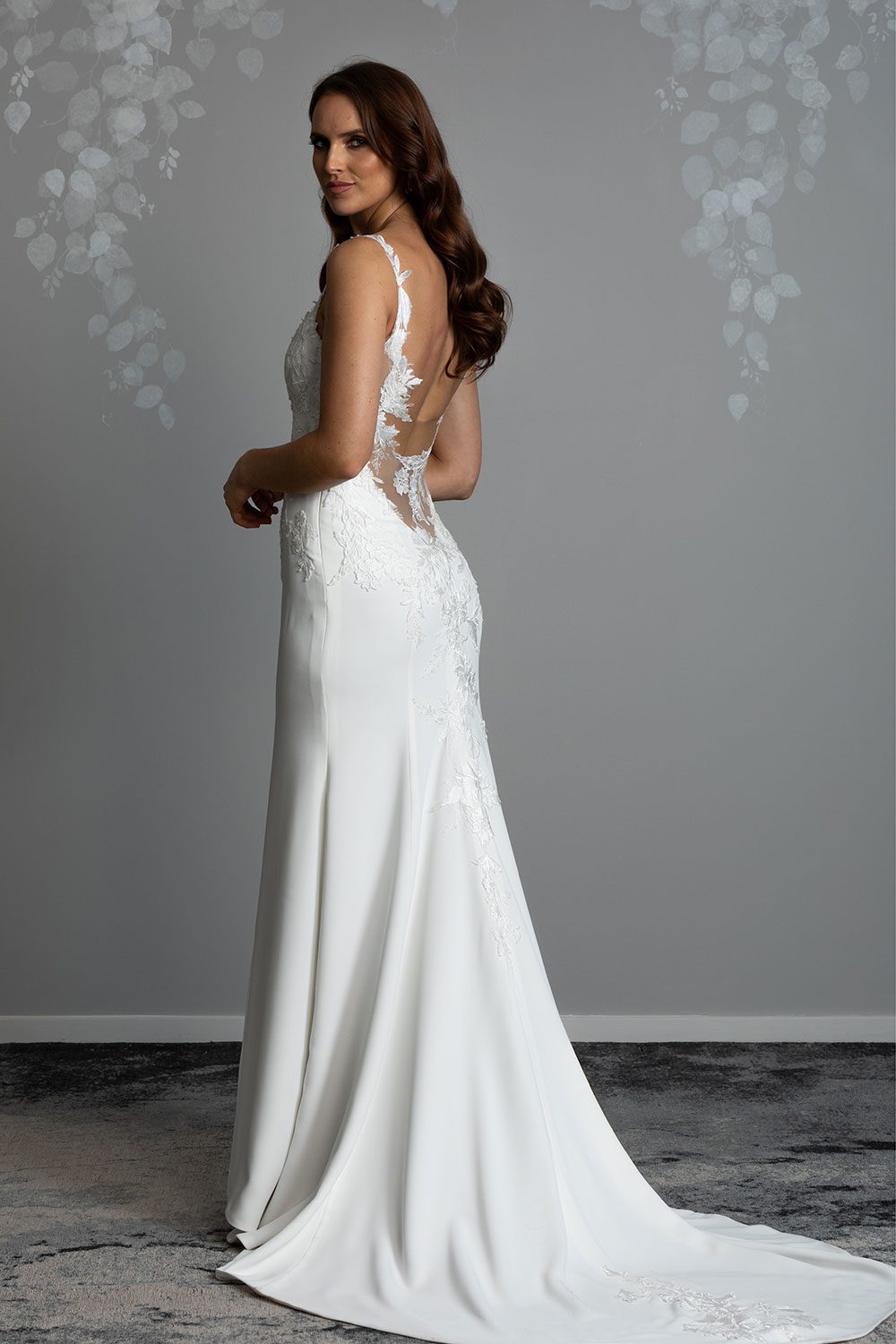 Ana Wedding gown from Vinka Design - This beautiful gown is graced by hand-appliqued delicate floral leaf lace, subtly worked into the front of the bodice and more prominently on the back. A fit-and-flare cut shapes the figure. Model wearing gown showing low back and lace detail - featured image