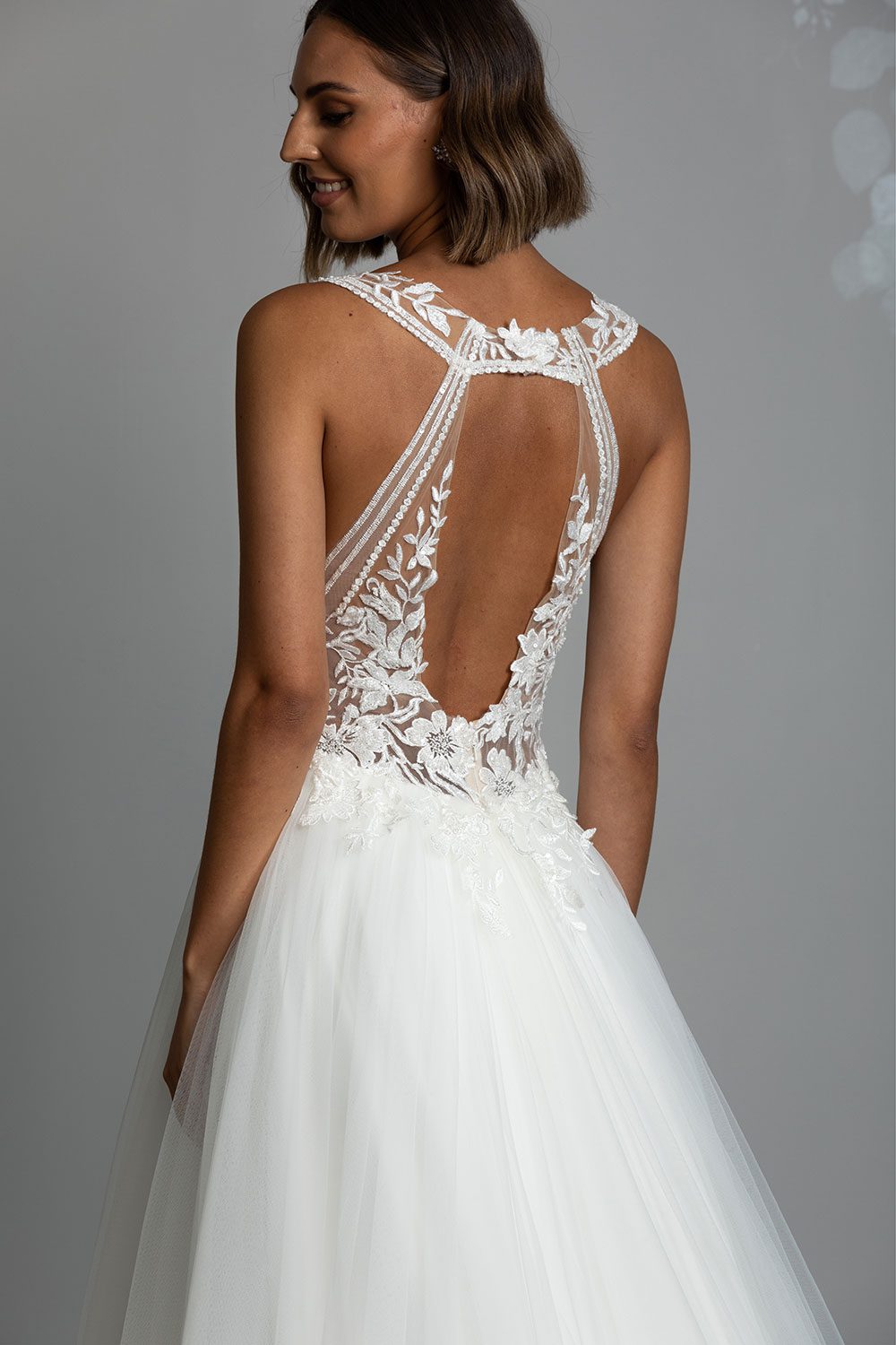 Tia Wedding gown from Vinka Design - This dreamy sculpted wedding dress has a deep V-shaped illusion neckline with beaded floral lace and an open back. The skirt has layers of soft tulle that glide with movement. Close up back view of dress with cut out floral lace open back