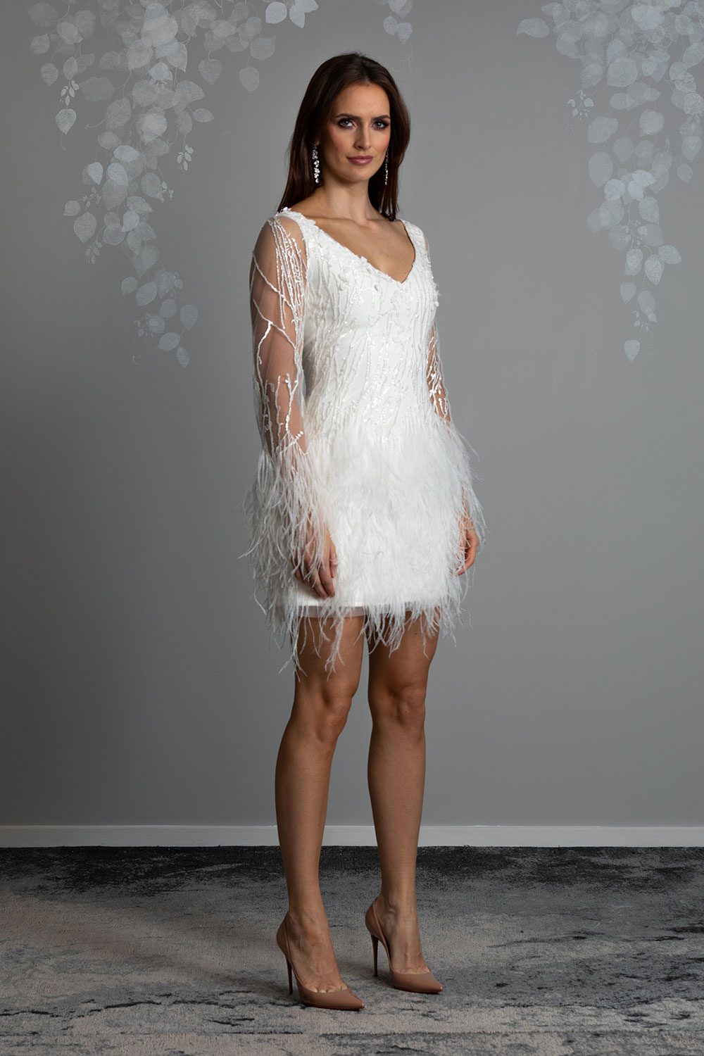 Nikora Wedding gown from Vinka Design - This sexy gown is sure to turn heads! Adorned with feathers that accentuate movement and bell sleeves that add flare, a high neckline, and low back. Semi profile view of model wearing mini dress with bell sleeves adorned with beads and feathers and V neckline