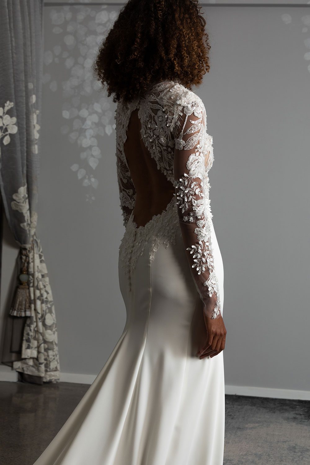 Moana Wedding gown from Vinka Design - Spectacular wedding dress perfect for the modern bride who still wants a classic spark! 3D lace embroidery complemented by a high neckline, fitted sleeves, and stunning low back into a flare train. Model showing close up of back of the gown with cut out 3D lace and embroidered back detail and long train