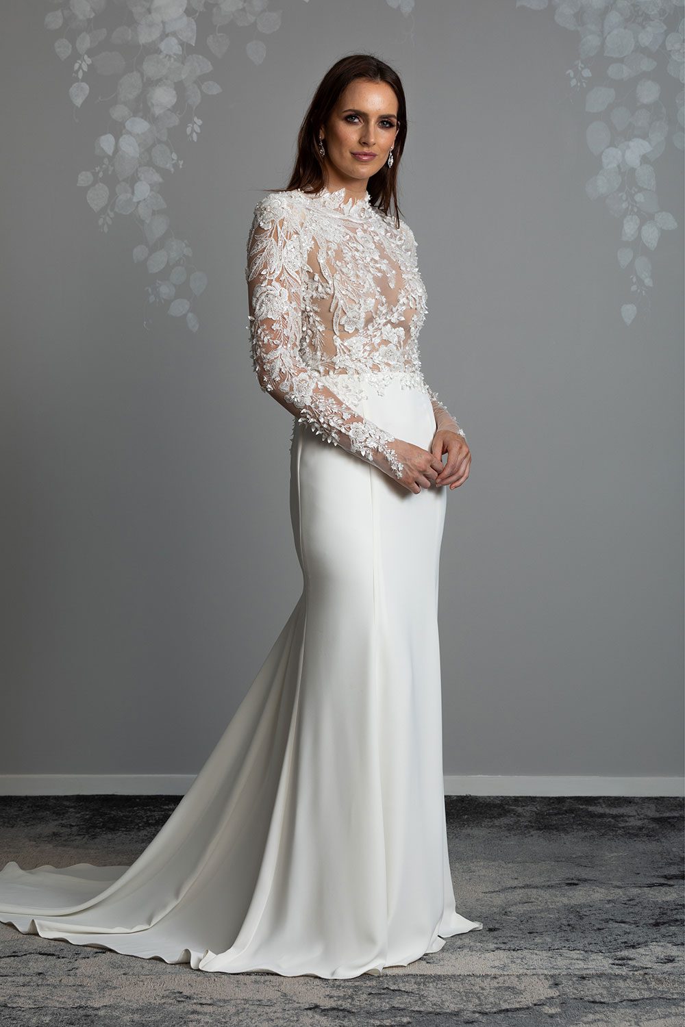 Moana Wedding gown from Vinka Design - Spectacular wedding dress perfect for the modern bride who still wants a classic spark! 3D lace embroidery complemented by a high neckline, fitted sleeves, and stunning low back into a flare train. Model with hands clasped in front wearing stunning classic high neck dress with long lace embroidered sleeves and fit and flare train