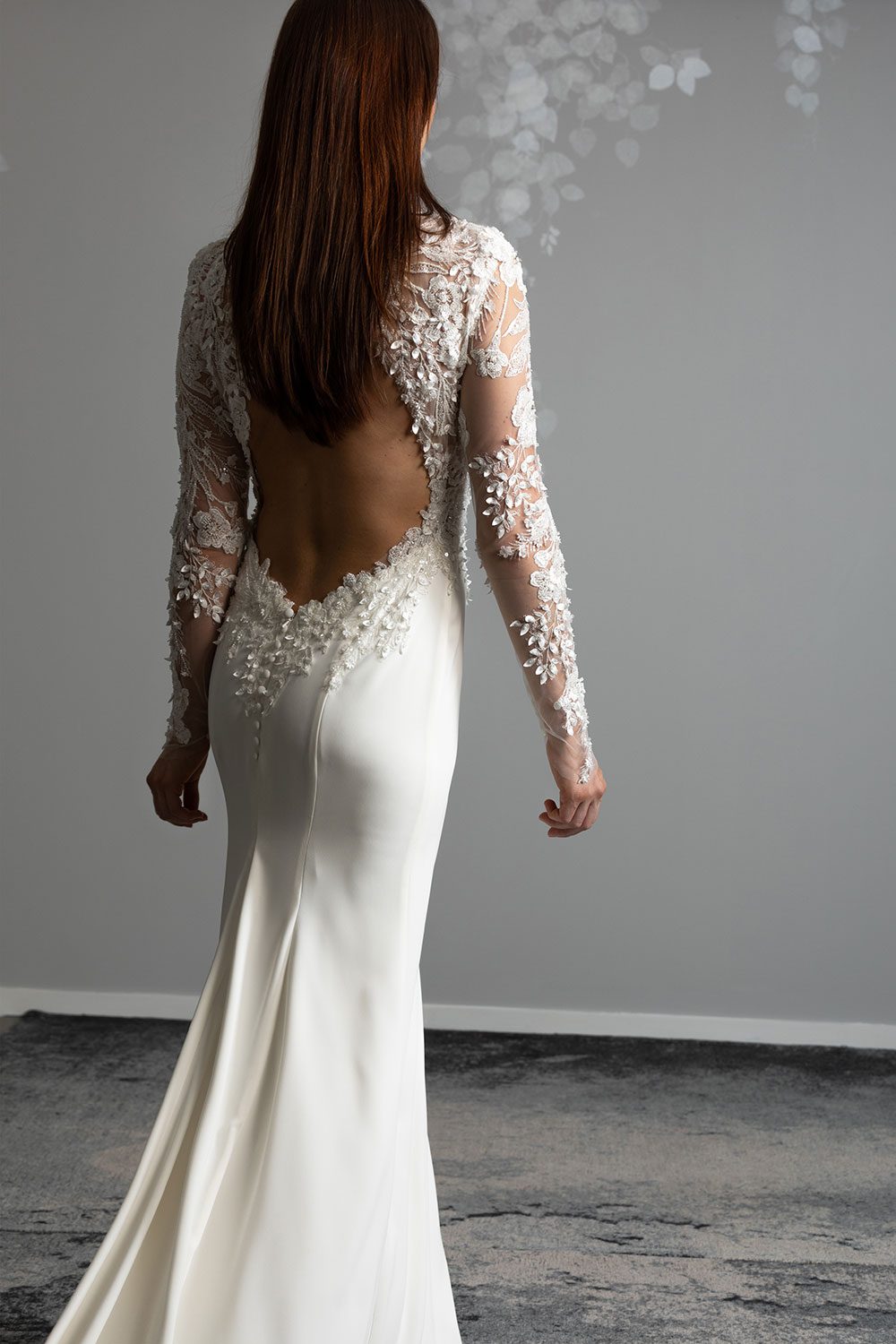 Moana Wedding gown from Vinka Design - Spectacular wedding dress perfect for the modern bride who still wants a classic spark! 3D lace embroidery complemented by a high neckline, fitted sleeves, and stunning low back into a flare train. Model showing back of the gown with cut out 3D lace and embroidered back detail and long train