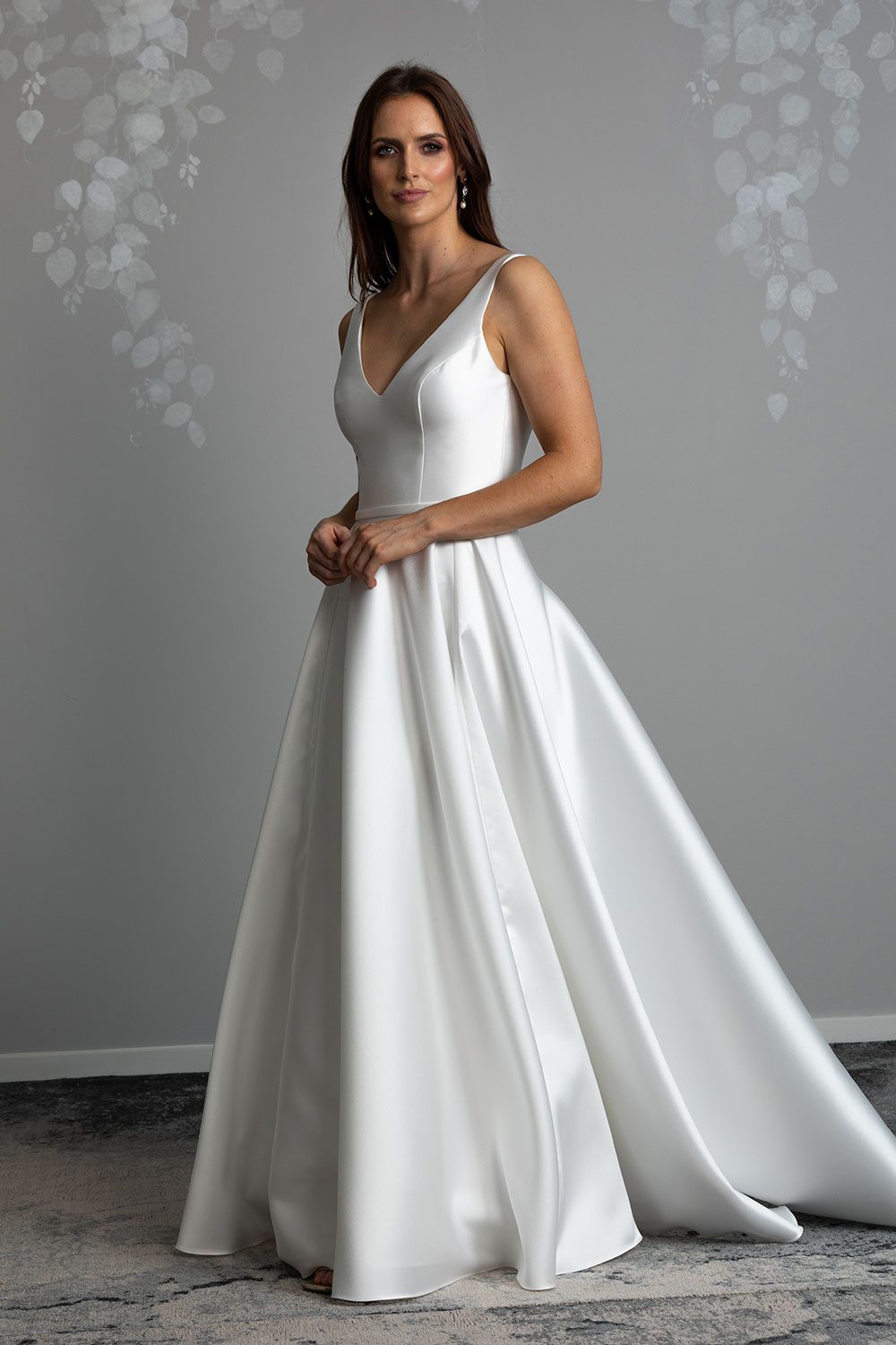 Madison Wedding gown from Vinka Design - This gown is both classic and contemporary, made beautifully with luxurious Mikado satin. The bodice is structured with a deep V-neckline and a squared, low back. Model with hands clasped across waist showing off the irregular folds in the skirt that fall elegantly into a long train