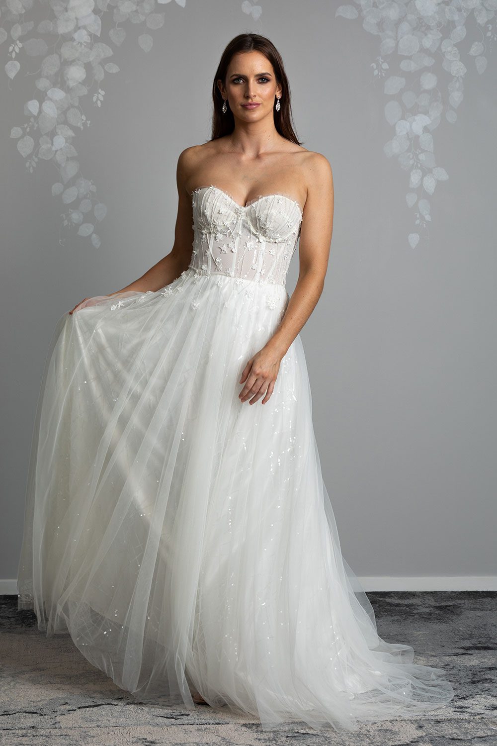 Hikari Wedding gown from Vinka Design - This modern wedding dress has a structured semi-sheer bodice with hand-appliqued lace of stars and flowers. The skirt is made with multiple layers of soft tulle. Full length image of model holding soft tulle skirt with sequined outer layer