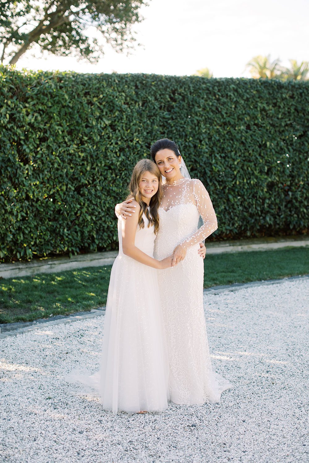 Bride wearing bespoke gown by Auckland wedding dress designer Vinka Design, with a high neckline and delicate spotted embroidery - with bridesmaid near hedge