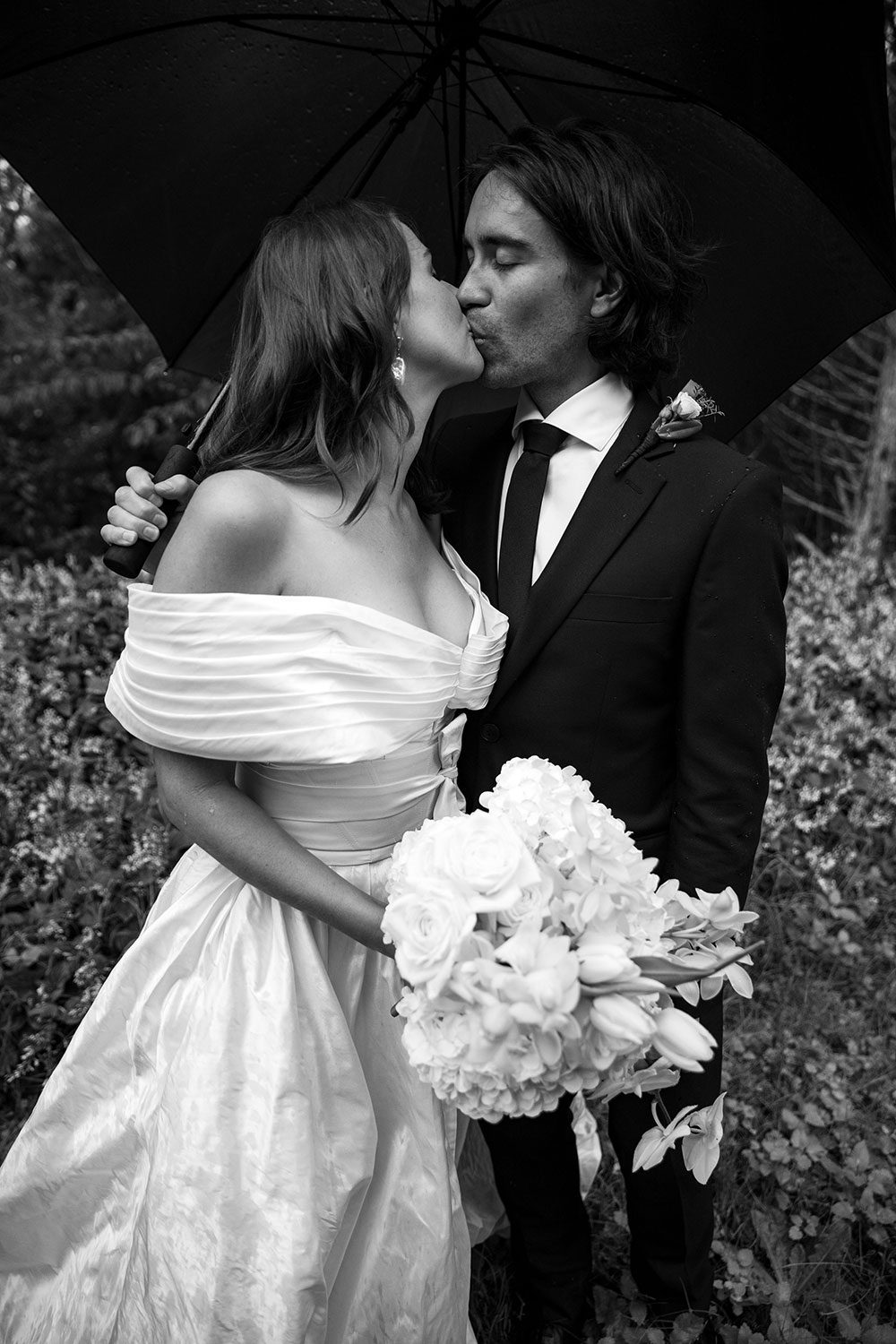 Bride wearing bespoke off shoulder bodice made with silk dupion and long train by NZ wedding dress maker Vinka Designs - black and white kissing groom under umbrella