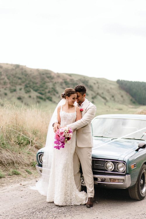 Bride wears bespoke E'More lace gown with boned bodice with hand beaded flower applique and full lace train by Auckland wedding dress maker Vinka designs - with groom in front of vintage car