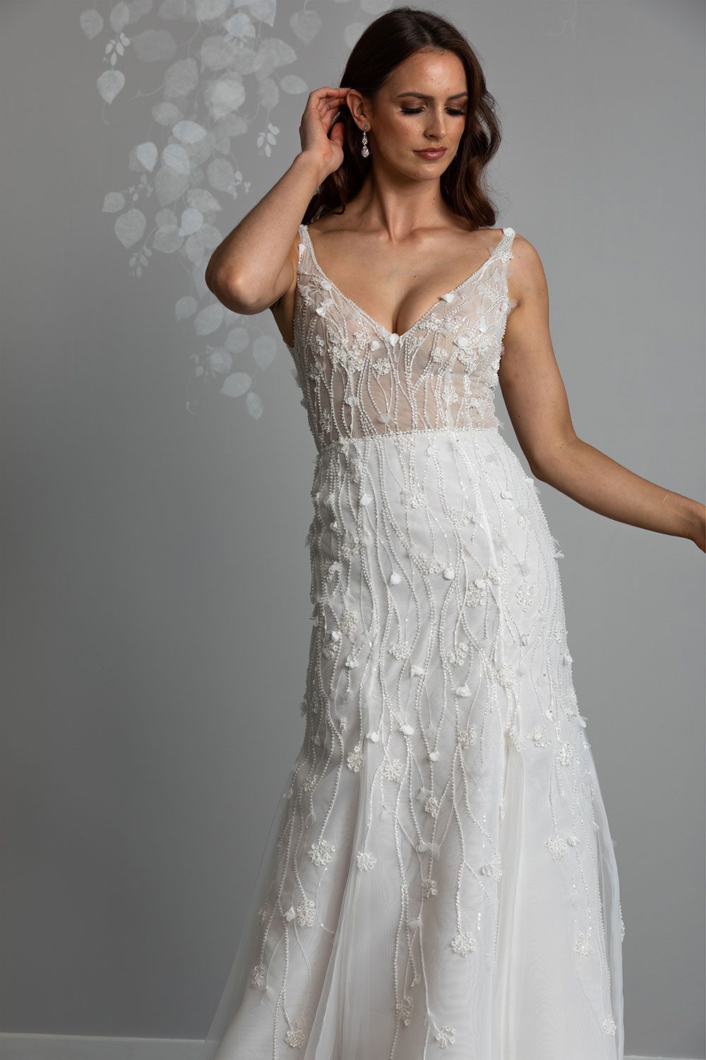Bronte Wedding Dress by Vinka Design - Featuring a form sculpting mermaid silhouette that embraces the wearer's curves, and intricately embroidered, beaded, botanical lace that trails seamlessly from the bodice into the skirt. Crafted on a semi-sheer base, the bodice boasts a V-neckline and beautiful low V-back. Artfully constructed with layers of dreamy, yet dramatic silk organza and soft tulle, added flare and fullness is achieved through the skirt which fans into a sweeping opulent train. Full length view of model with hand to hair wearing V-neckline bodice with beaded botanical lace with a dreamy silk organza and soft tulle skirt