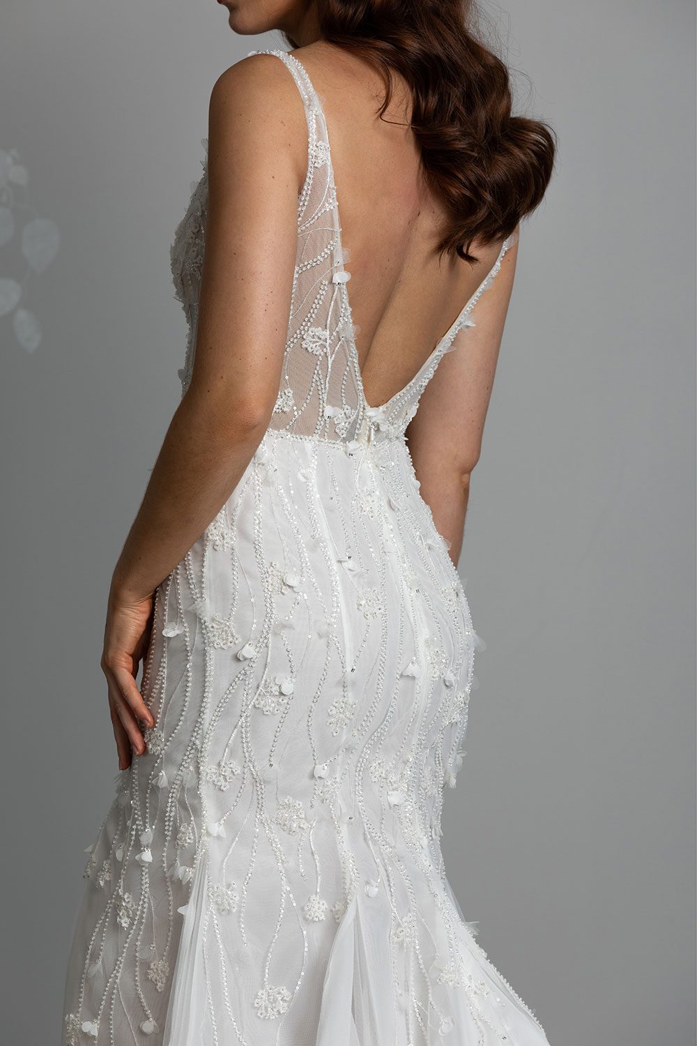Bronte Wedding Dress by Vinka Design - Featuring a form sculpting mermaid silhouette that embraces the wearer's curves, and intricately embroidered, beaded, botanical lace that trails seamlessly from the bodice into the skirt. Crafted on a semi-sheer base, the bodice boasts a V-neckline and beautiful low V-back. Artfully constructed with layers of dreamy, yet dramatic silk organza and soft tulle, added flare and fullness is achieved through the skirt which fans into a sweeping opulent train. Back view of model wearing low V back bodice with intricately embroidered beaded botanical lace