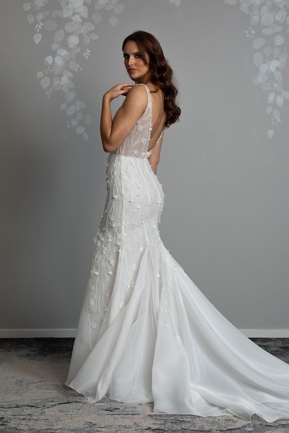 Bronte Wedding Dress by Vinka Design - Featuring a form sculpting mermaid silhouette that embraces the wearer's curves, and intricately embroidered, beaded, botanical lace that trails seamlessly from the bodice into the skirt. Crafted on a semi-sheer base, the bodice boasts a V-neckline and beautiful low V-back. Artfully constructed with layers of dreamy, yet dramatic silk organza and soft tulle, added flare and fullness is achieved through the skirt which fans into a sweeping opulent train. Profile view of model with hand to shoulder wearing low V back bodice with intricately embroidered beaded botanical lace and full skirt with sweeping train