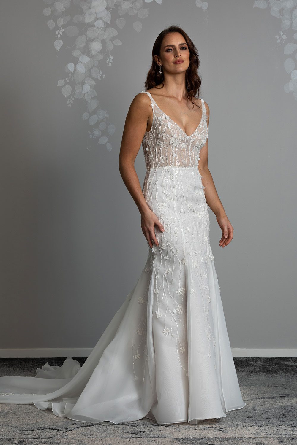 Bronte Wedding Dress by Vinka Design - Featuring a form sculpting mermaid silhouette that embraces the wearer's curves, and intricately embroidered, beaded, botanical lace that trails seamlessly from the bodice into the skirt. Crafted on a semi-sheer base, the bodice boasts a V-neckline and beautiful low V-back. Artfully constructed with layers of dreamy, yet dramatic silk organza and soft tulle, added flare and fullness is achieved through the skirt which fans into a sweeping opulent train. Full length view of model wearing V-neckline bodice with intricately embroidered beaded botanical lace and full skirt with sweeping train