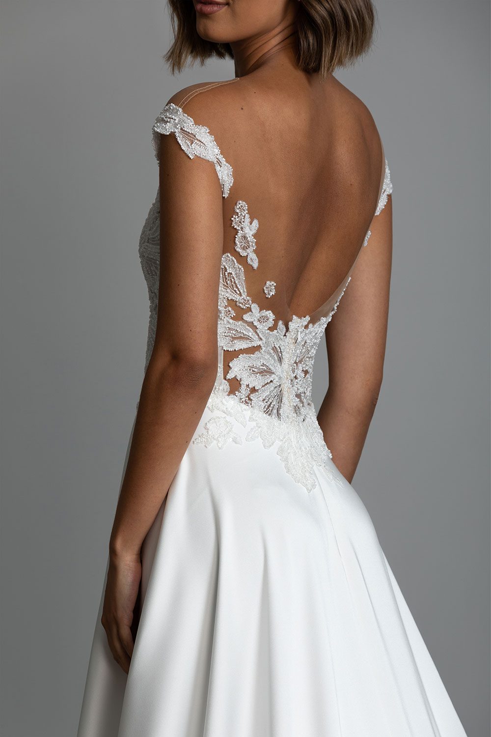 Charlotte Wedding Gown by Vinka Design - Featuring a delicate sweetheart neckline and off shoulder sleeves that draw the eye to the wearer’s décolletage. Botanical, intricately beaded, embroidered lace graces the fitted, boned bodice and trails off the shoulder into a low back on a nude, illusion tulle base. Close up of back of model wearing off shoulder sleeves of beaded botanical lace and sweetheart neckline with A-line skirt and cascading train