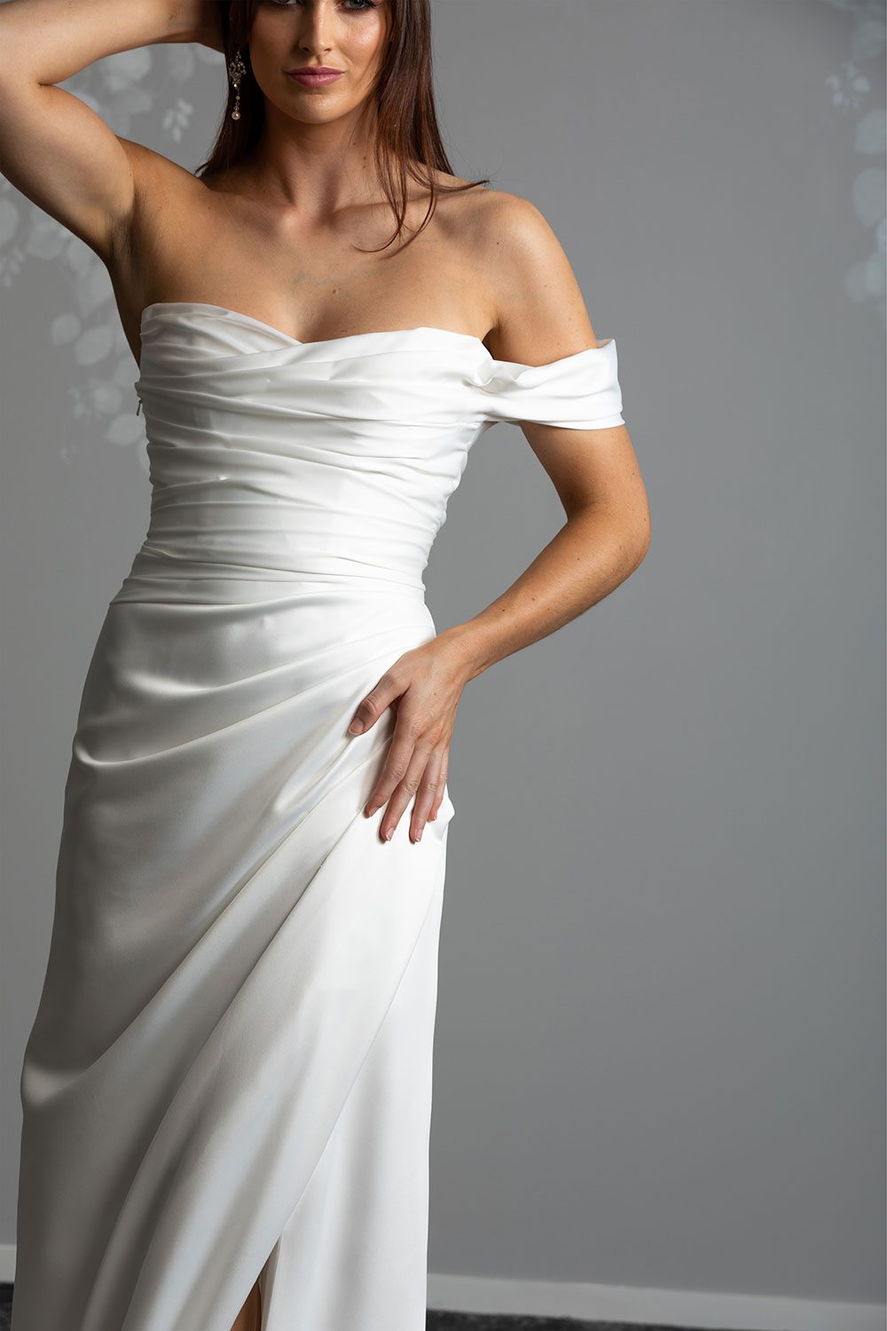 Christina Wedding Dress by Vinka Design - Featuring a sculptured, fitted bodice with a scoop neckline that flows seamlessly into a beautiful off shoulder detail. The draping from the bodice drops from the hips into a gentle fit and flare skirt, falling into an elegant train and features a concealed side split that reveals a subtle hint of leg. Close up of model showing off one shoulder detail and draped fitted bodice with scoop neckline
