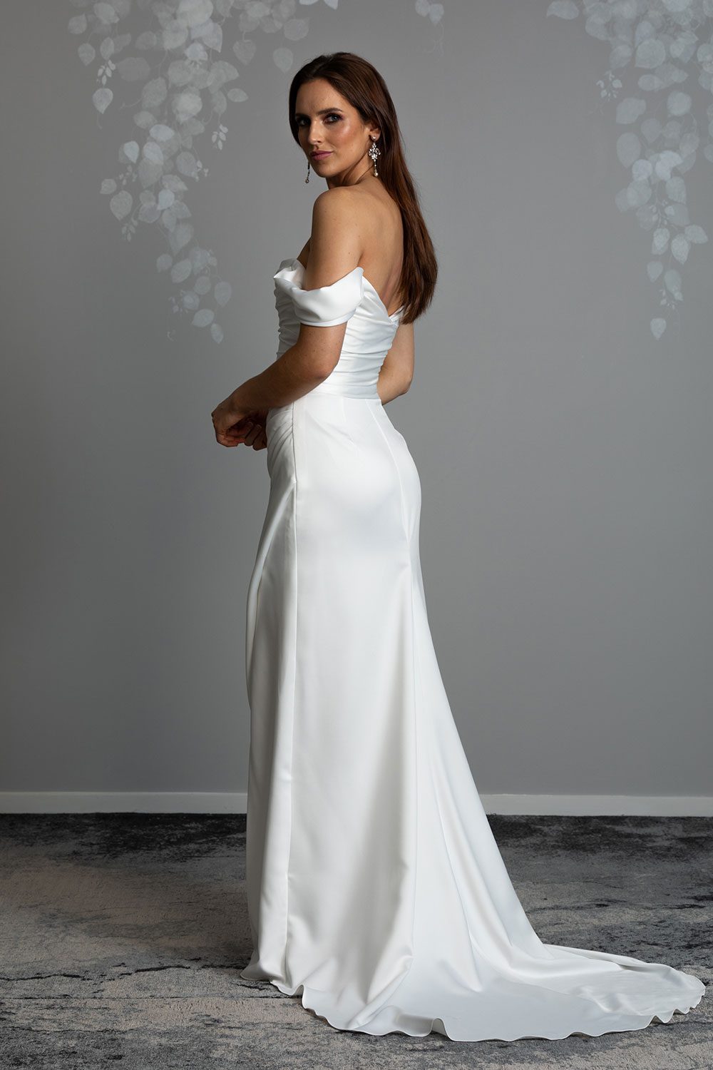 Christina Wedding Dress by Vinka Design - Featuring a sculptured, fitted bodice with a scoop neckline that flows seamlessly into a beautiful off shoulder detail. The draping from the bodice drops from the hips into a gentle fit and flare skirt, falling into an elegant train and features a concealed side split that reveals a subtle hint of leg. Profile view of model showing off shoulder detail and fitted bodice with fit and flare skirt with train