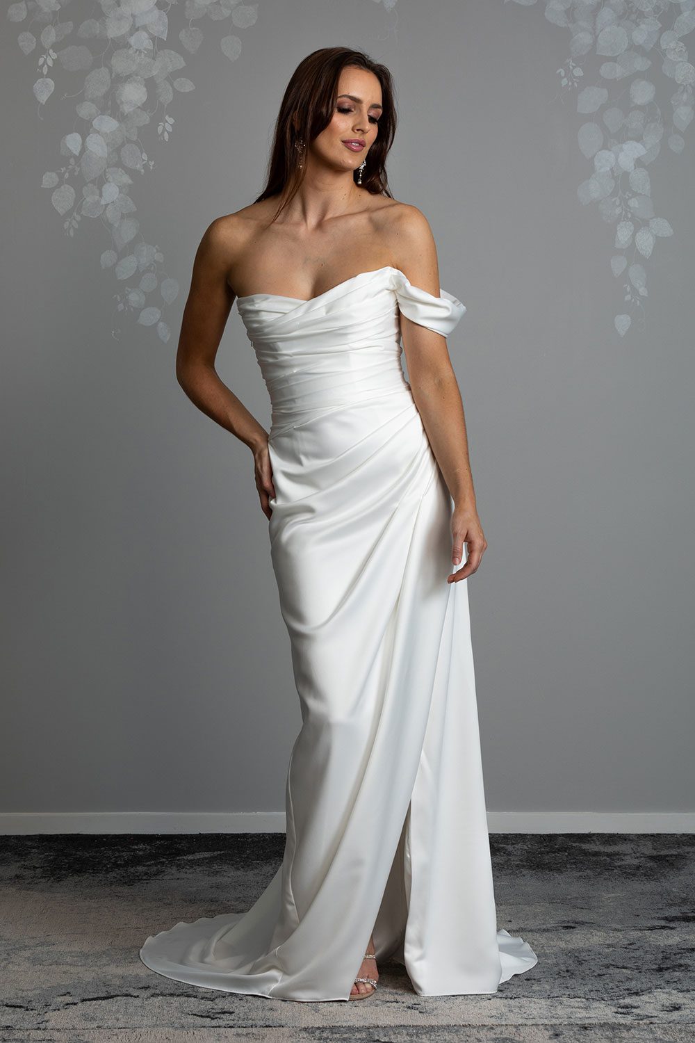 Christina Wedding Dress by Vinka Design - Featuring a sculptured, fitted bodice with a scoop neckline that flows seamlessly into a beautiful off shoulder detail. The draping from the bodice drops from the hips into a gentle fit and flare skirt, falling into an elegant train and features a concealed side split that reveals a subtle hint of leg. Model looking over shoulder with hand to hip wearing delicately draped silky satin dress over a fitted bodice and one shoulder detail