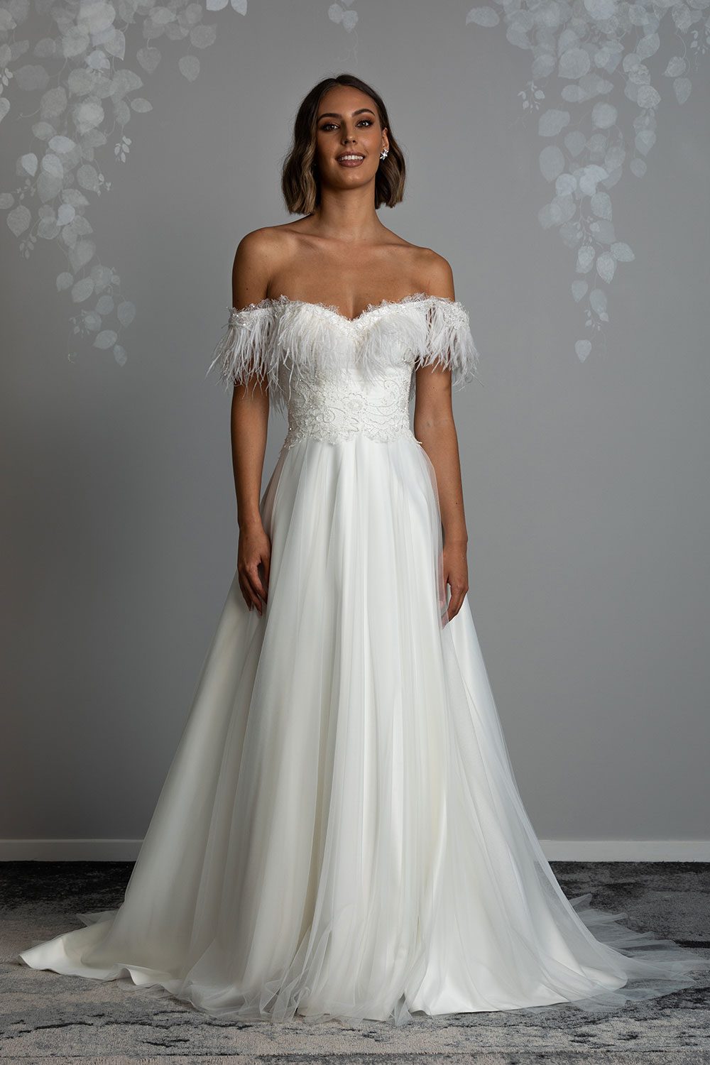 Heather wedding Dress by Vinka Design - Featuring a classic structured bodice hand-appliquéd with intricately beaded french lace accentuating the waist, which spills into a skirt crafted with full feather-light tulle. The off-shoulder bodice draws the eye to the wearer’s décolletage and is adorned opulently with ostrich feathers and appliquéd with iridescent beaded detailing. Front view of model wearing dreamy feathered structured bodice gown with skirt made of layers of tulle