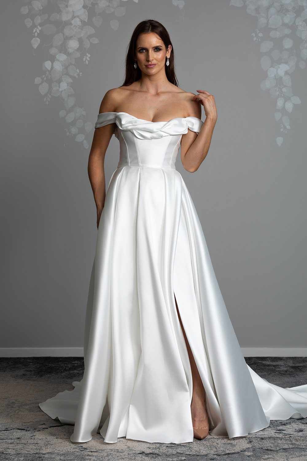 Helena Wedding Dress by Vinka Design - . The statement scoop neckline and daring cross-over concealed leg split featured on our Helena gown is complimented with a nostalgic ball gown silhouette. The classic corseted bodice is adorned with a draped off-shoulder detail, framing the neckline whilst adding definition and drawing the eye to the wearer’s waist which spills into luxe layers of Japanese Mikado satin. Full length view of model with hand to shoulder showing of draped off shoulder detailing of neckline