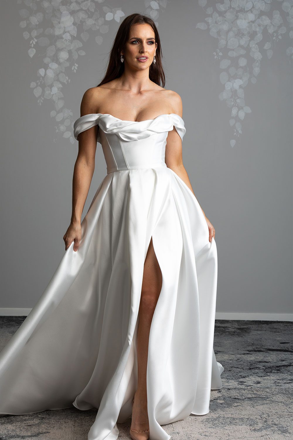 Helena Wedding Dress by Vinka Design - The statement scoop neckline and daring cross-over concealed leg split featured on our Helena gown is complimented with a nostalgic ball gown silhouette. The classic corseted bodice is adorned with a draped off-shoulder detail, framing the neckline whilst adding definition and drawing the eye to the wearer’s waist which spills into luxe layers of Japanese Mikado satin. Model walking forward showing off front split