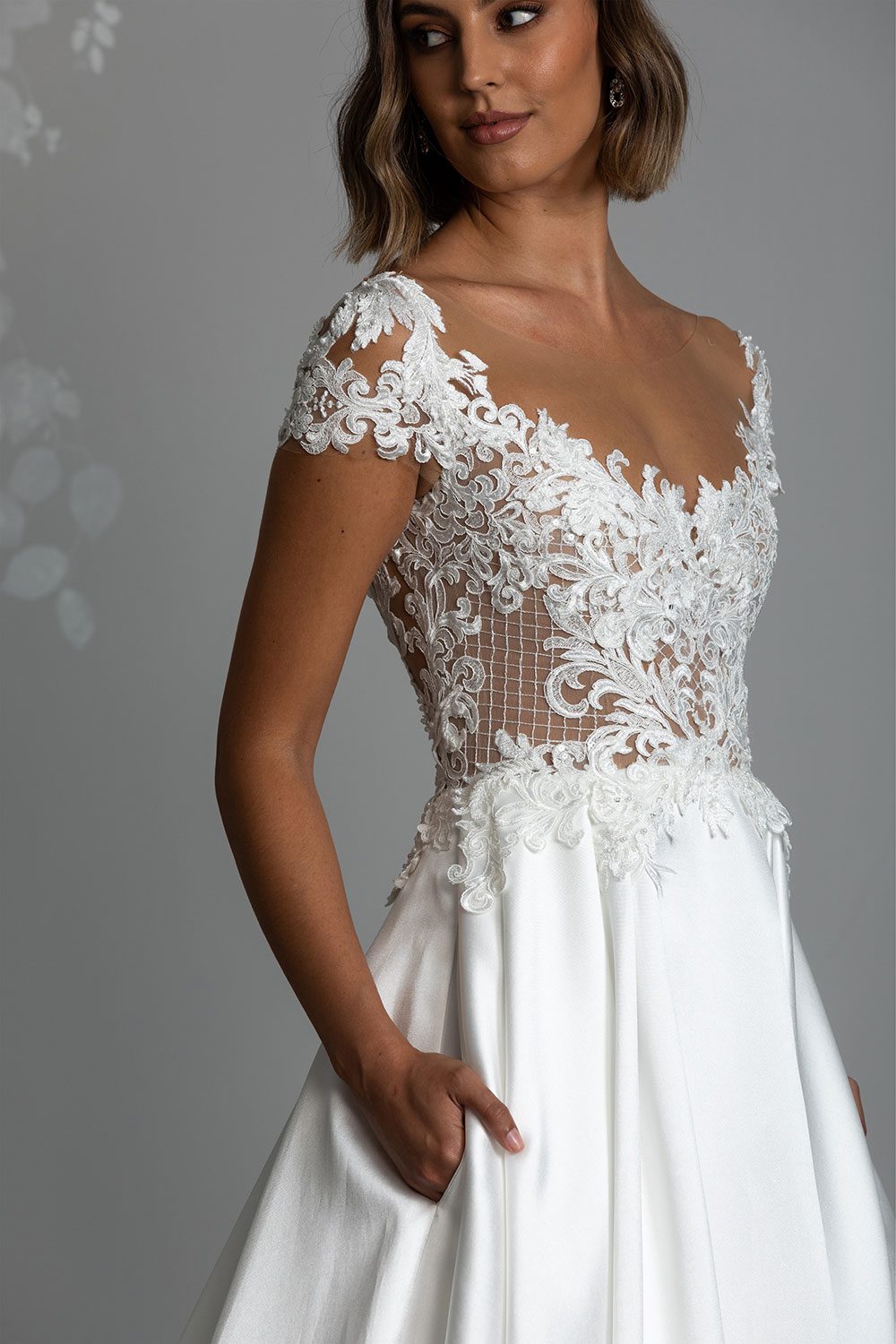 Leona Wedding Dress from Vinka Design. This gorgeous gown is hand appliqued with two complimentary laces on a nude tulle base, which creates a striking effect. Delicate cap sleeves flatter the arms whilst also drawing the eye to the slenderness of the waist. It has a low back edged with lace that cinches in the waist and is carefully styled to delicately finish into the dramatic skirt. Close up of front view of model showing long intricate lace bodice with cap sleeves and silk skirt with deep folds and pockets
