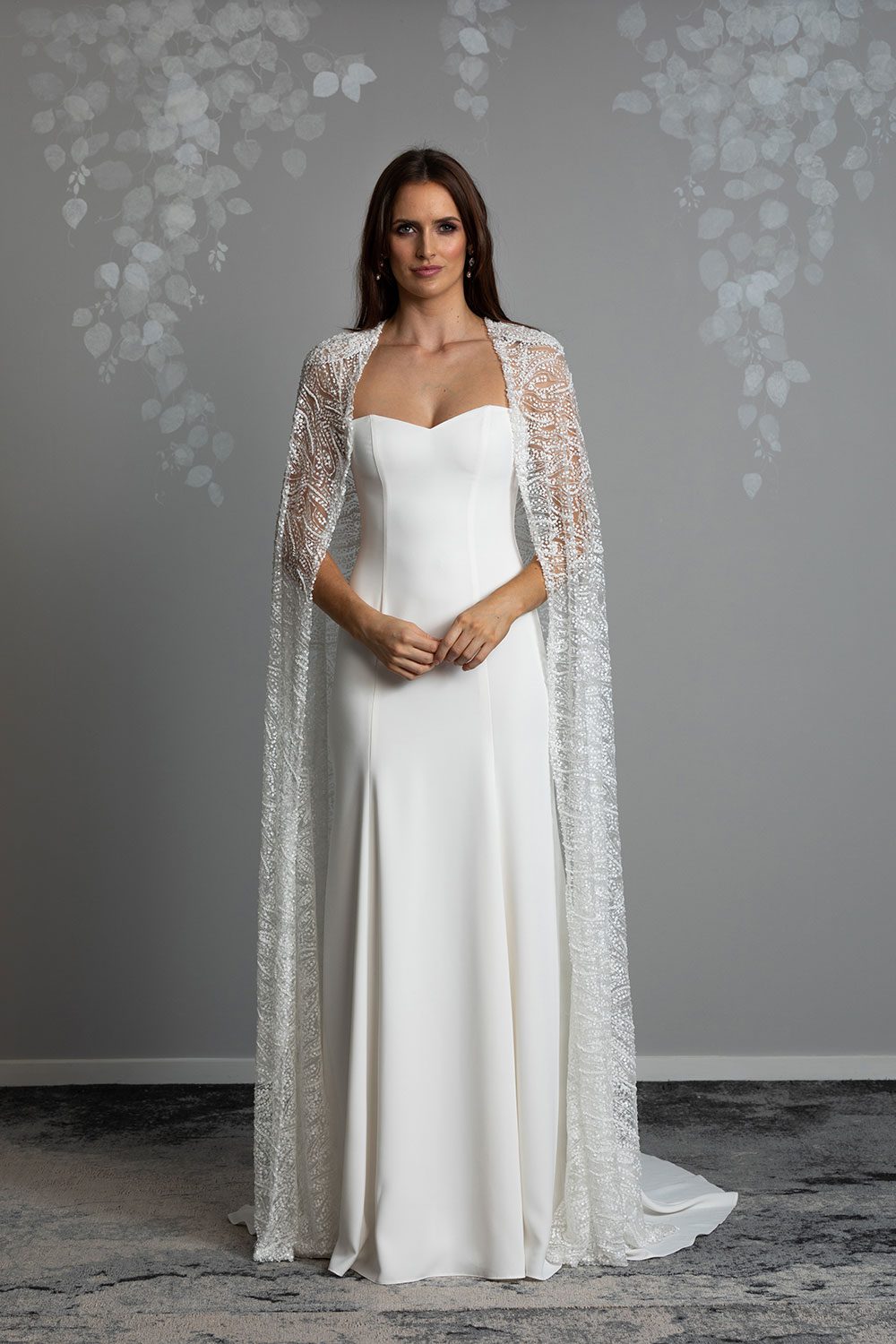 Lourde wedding Dress by Vinka Design - A dazzling display of beaded lace draped artfully from the shoulder, our Lourde gown is the epitome of timeless bridal elegance. Lourde’s cape features accentuated shoulders and opulent, iridescent beading. Crafted with functionality in mind, the richly beaded cape is detachable and reveals a panel cut gown with a fit-and-flare silhouette made in luxurious bridal crepe that falls flawlessly into a flared train. Full length view of model wearing sweetheart neckline gown with thin straps made of bridal crepe with a beaded cape and flared train
