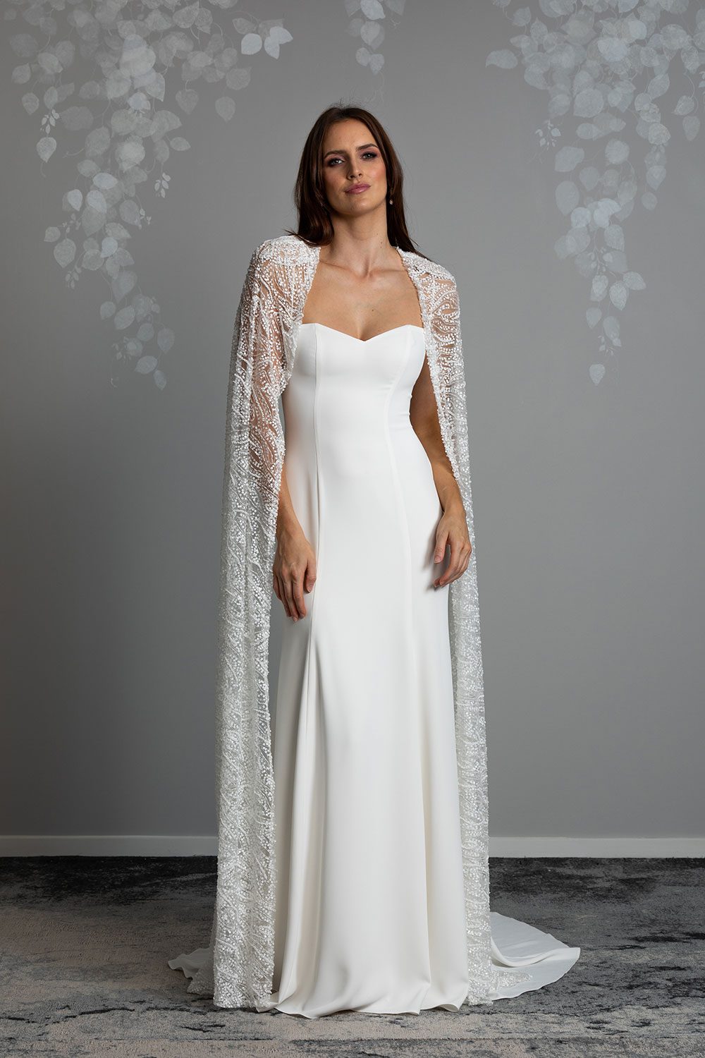 Lourde wedding Dress by Vinka Design - A dazzling display of beaded lace draped artfully from the shoulder, our Lourde gown is the epitome of timeless bridal elegance. Lourde’s cape features accentuated shoulders and opulent, iridescent beading. Crafted with functionality in mind, the richly beaded cape is detachable and reveals a panel cut gown with a fit-and-flare silhouette made in luxurious bridal crepe that falls flawlessly into a flared train. Full length view of model wearing sweetheart neckline gown with thin straps made of bridal crepe with a beaded cape draped over shoulders and flared train