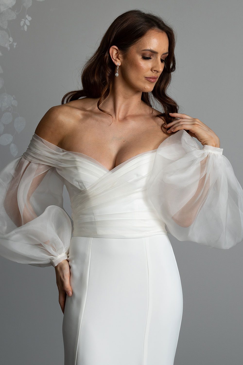 Marilyn Wedding Dress by Vinka Design - . Featuring a sculptured, fitted bodice with a sweetheart neckline that flows seamlessly into beautiful off shoulder detail that dips into a V-shaped back. For added drama, we have paired our Marilyn gown with detachable silk organza sleeves that taper at the wrist with delicate, self-covered silk buttons. Model with hand to shoulder and looking down wearing organza detachable statement sleeves over timeless design gown
