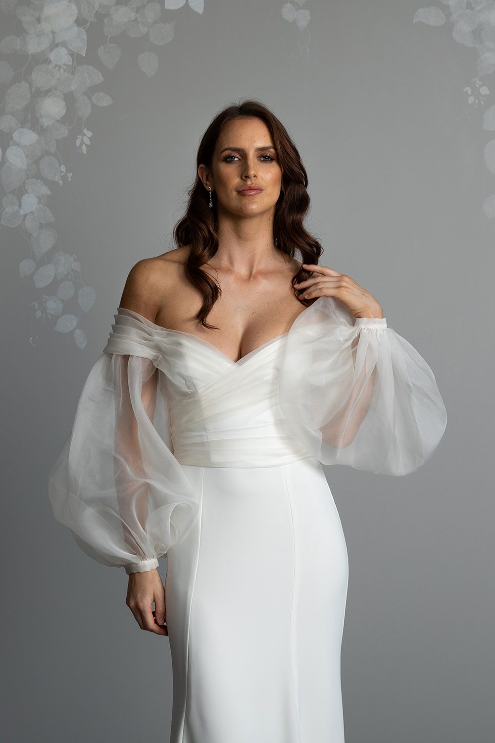 Marilyn Wedding Dress by Vinka Design - . Featuring a sculptured, fitted bodice with a sweetheart neckline that flows seamlessly into beautiful off shoulder detail that dips into a V-shaped back. For added drama, we have paired our Marilyn gown with detachable silk organza sleeves that taper at the wrist with delicate, self-covered silk buttons. Model with hand to shoulder and looking forward wearing organza detachable statement sleeves over timeless design gown