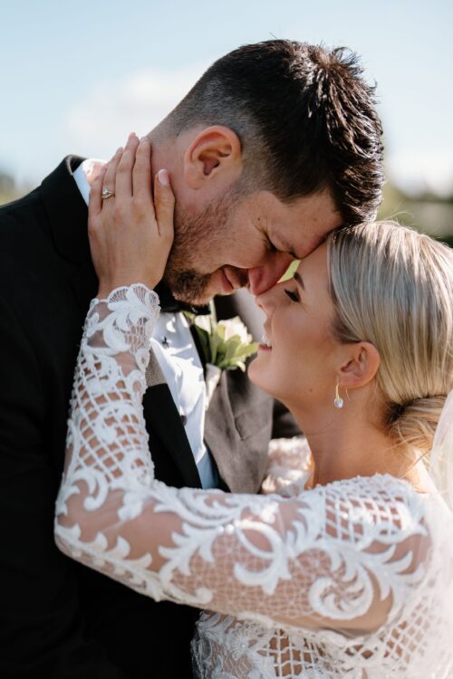 Groom and bride wearing Vinka Design bespoke gown, featuring a high neckline, intricate beading, long, lace sleeves, in a luxurious bridal crepe base embrace