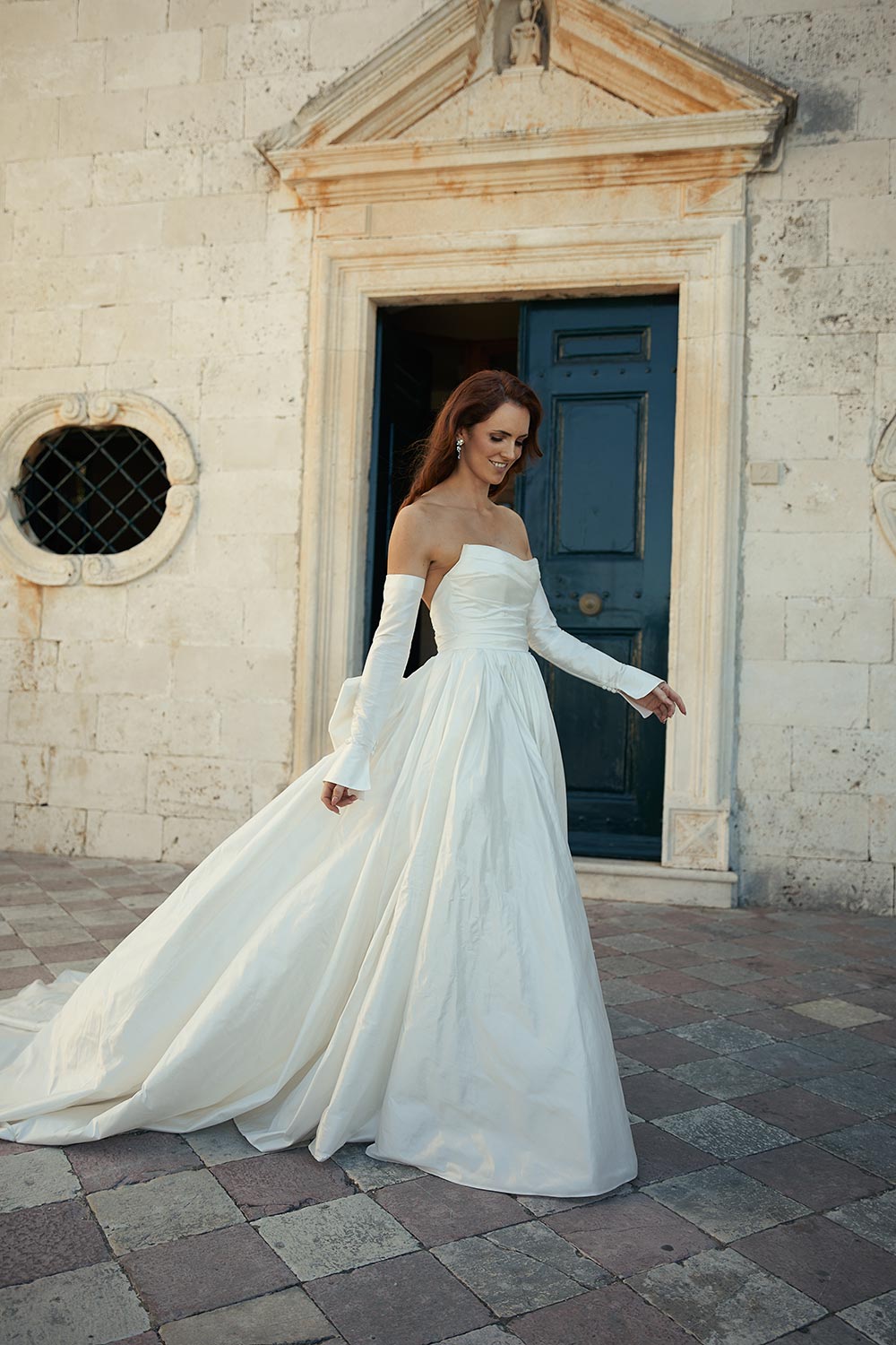 Discover Marija, a layered dupion silk wedding gown of regal romance. Features a scoop neckline, draped bodice, detachable sleeves and bow, and a long train. By Vinka Design